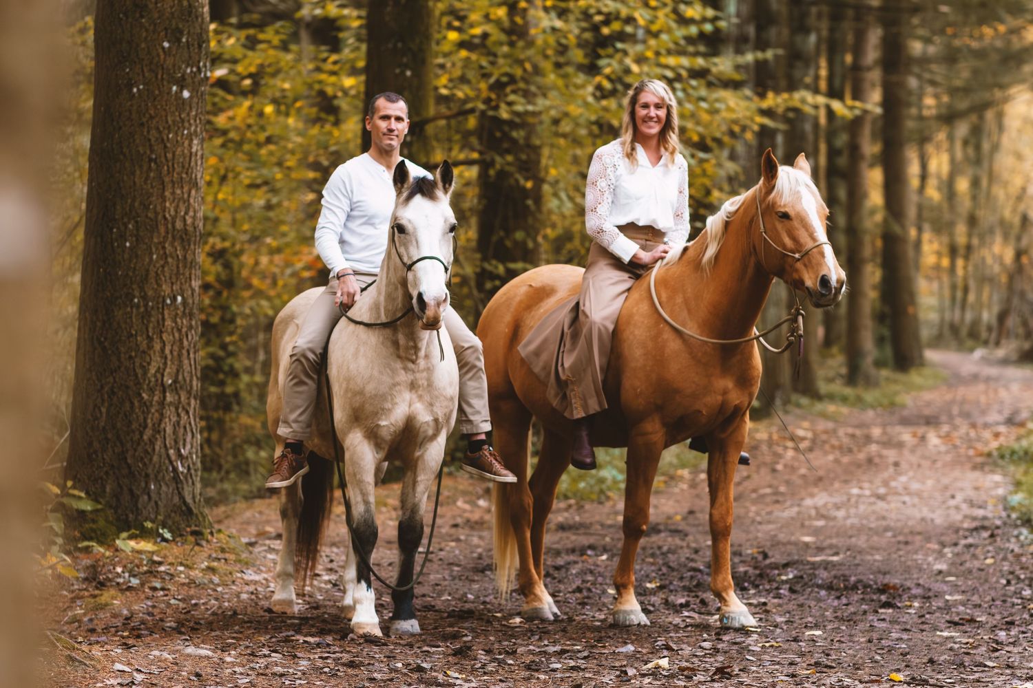 Couple riding chestnut and grey horses side by side on a scenic autumn forest path.