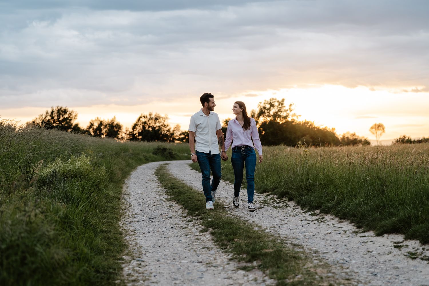 Couple holding hands walking on a winding dirt path through green fields at golden sunset.
