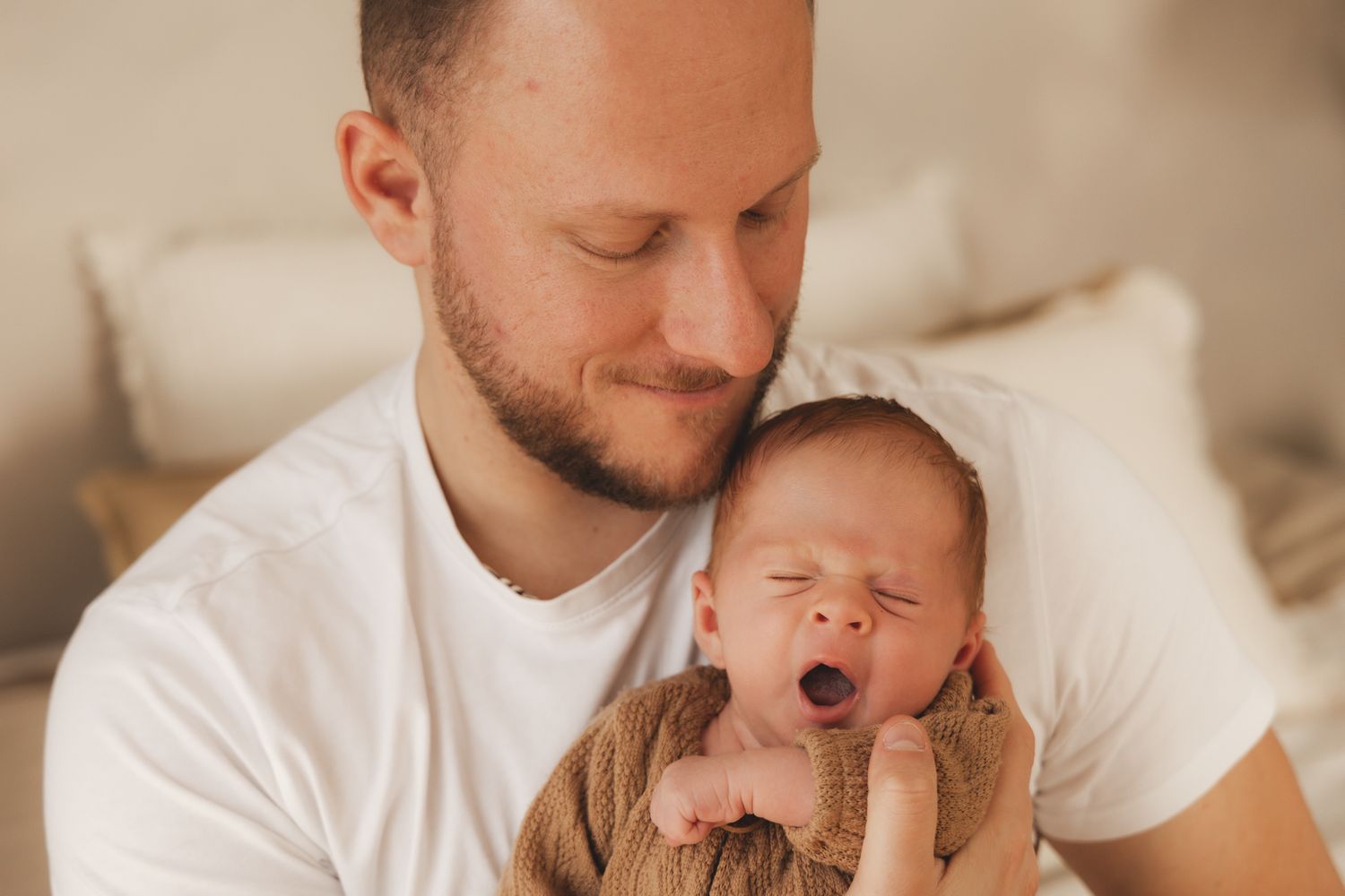 Father in white t-shirt holding and looking lovingly at his yawning newborn baby.