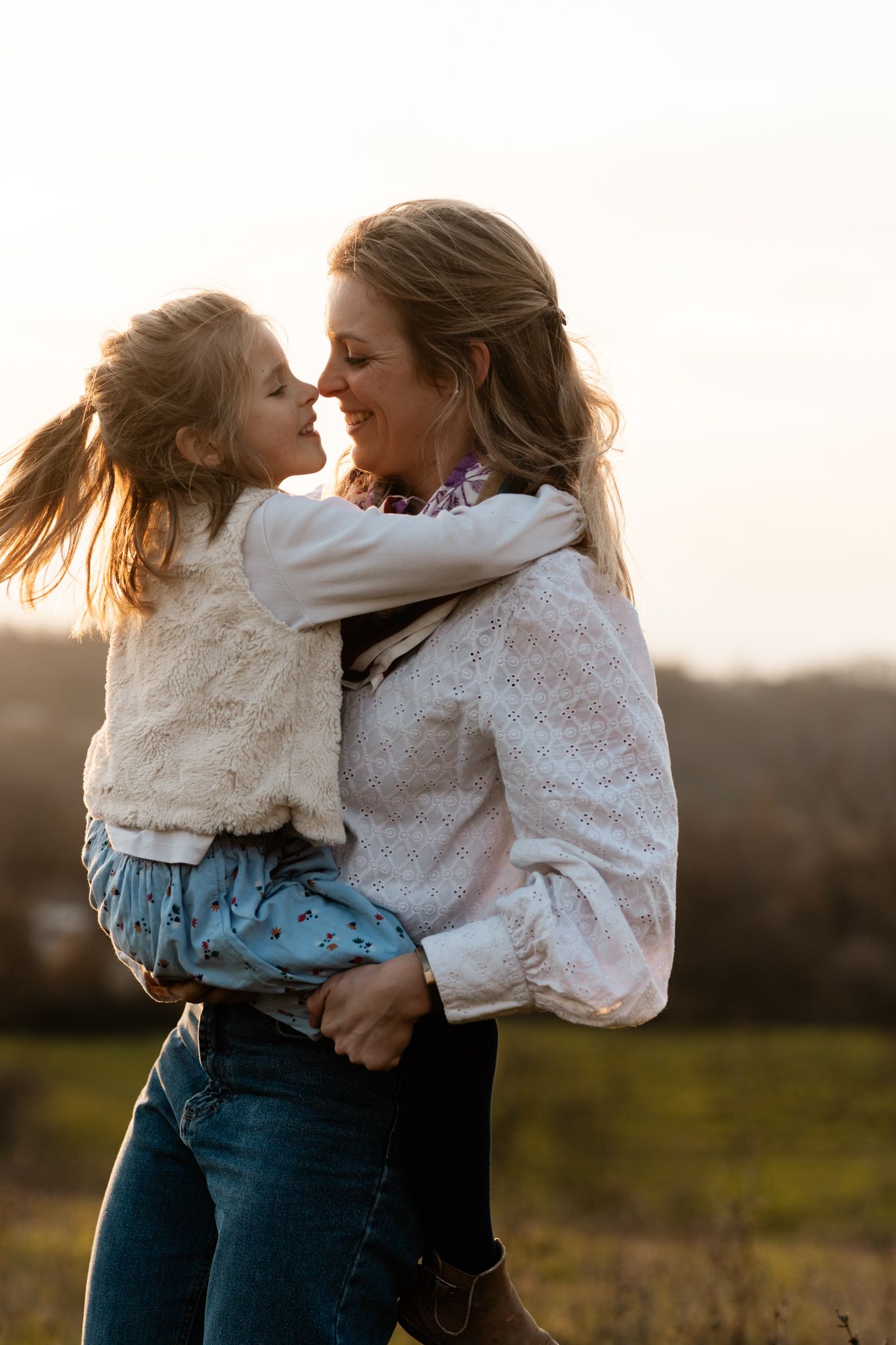 Séance photo famille en Isère : une mère souriante tient sa petite fille face à face à la campagne au coucher du soleil.