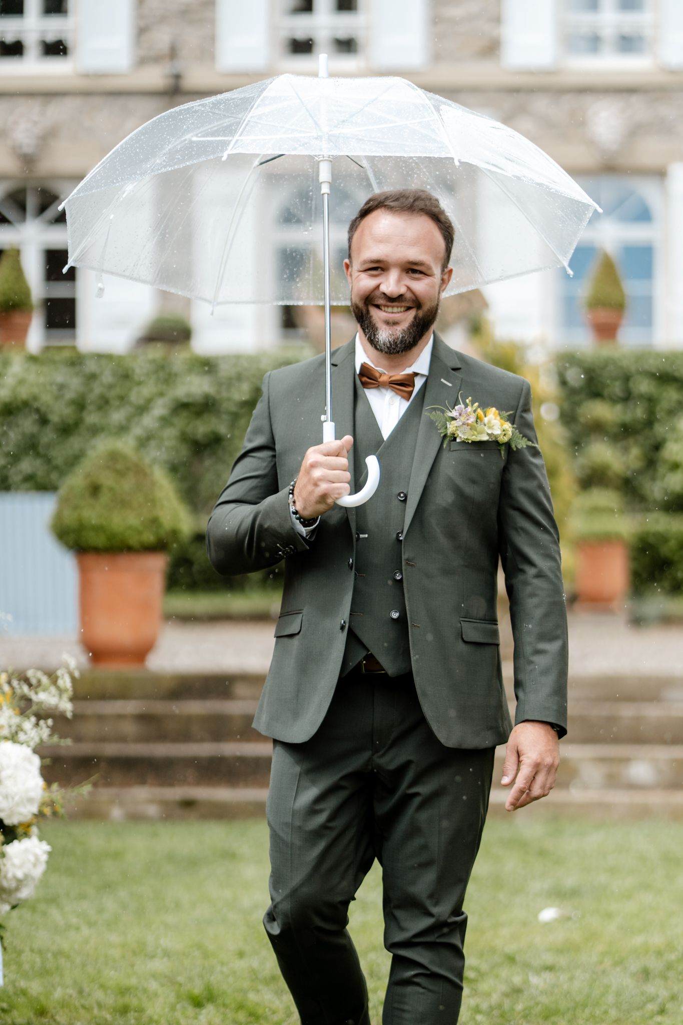 Smiling groom in dark green suit holding clear umbrella with boutonniere at outdoor wedding ceremony.