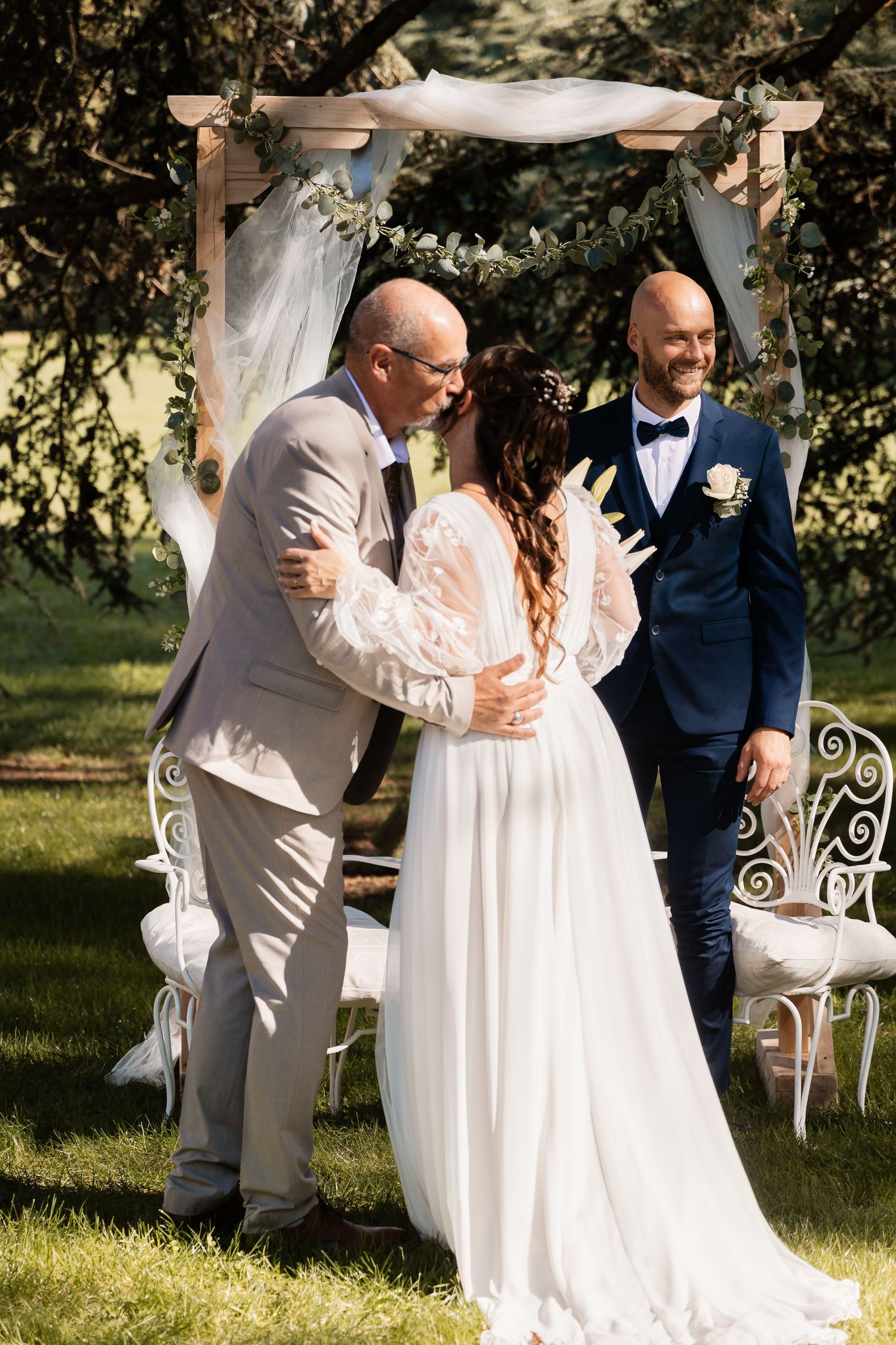 Bride and groom share their first kiss at an outdoor wedding ceremony beneath a floral arch.