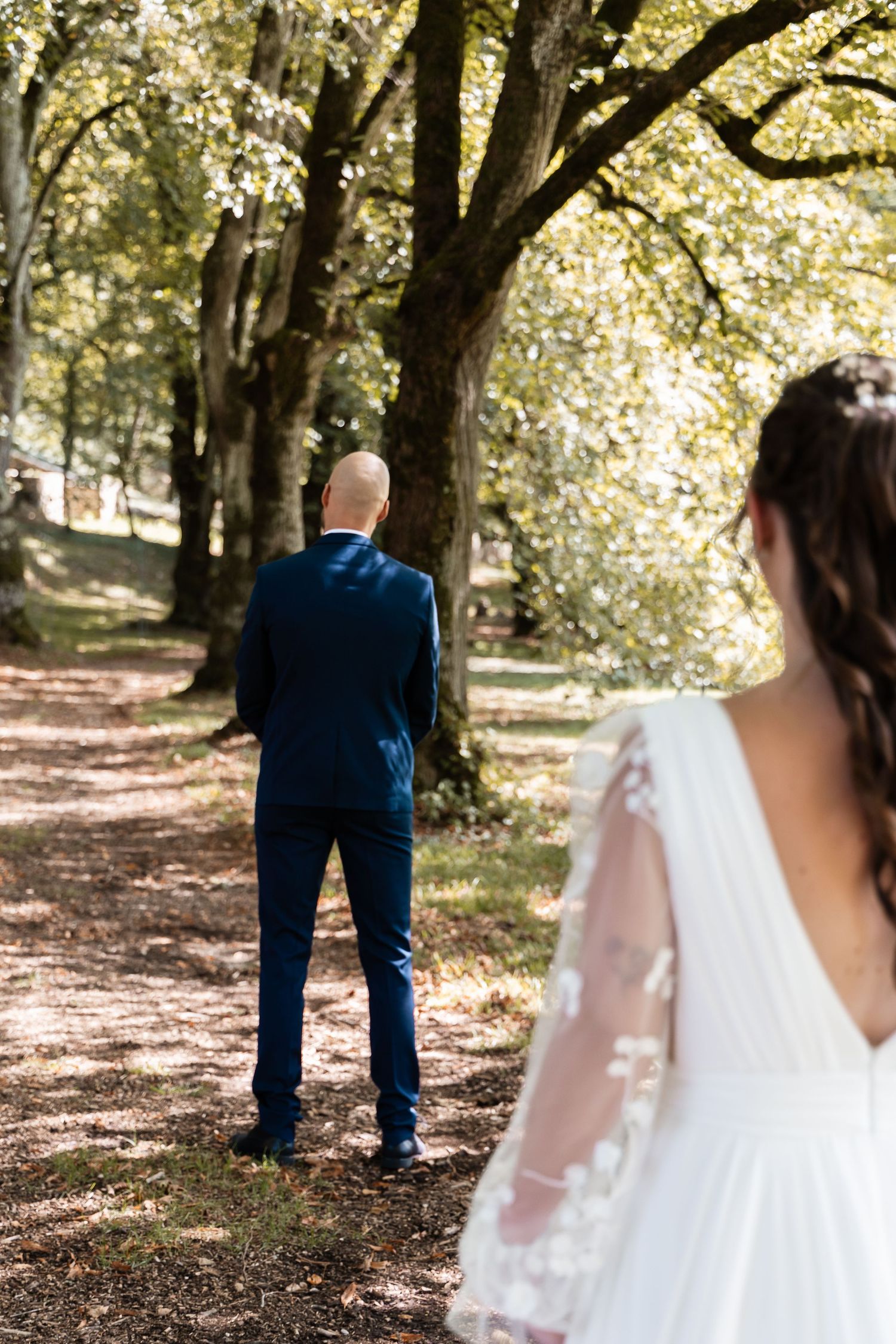Groom in navy suit stands with back turned on forest path as bride in lace wedding dress approaches.