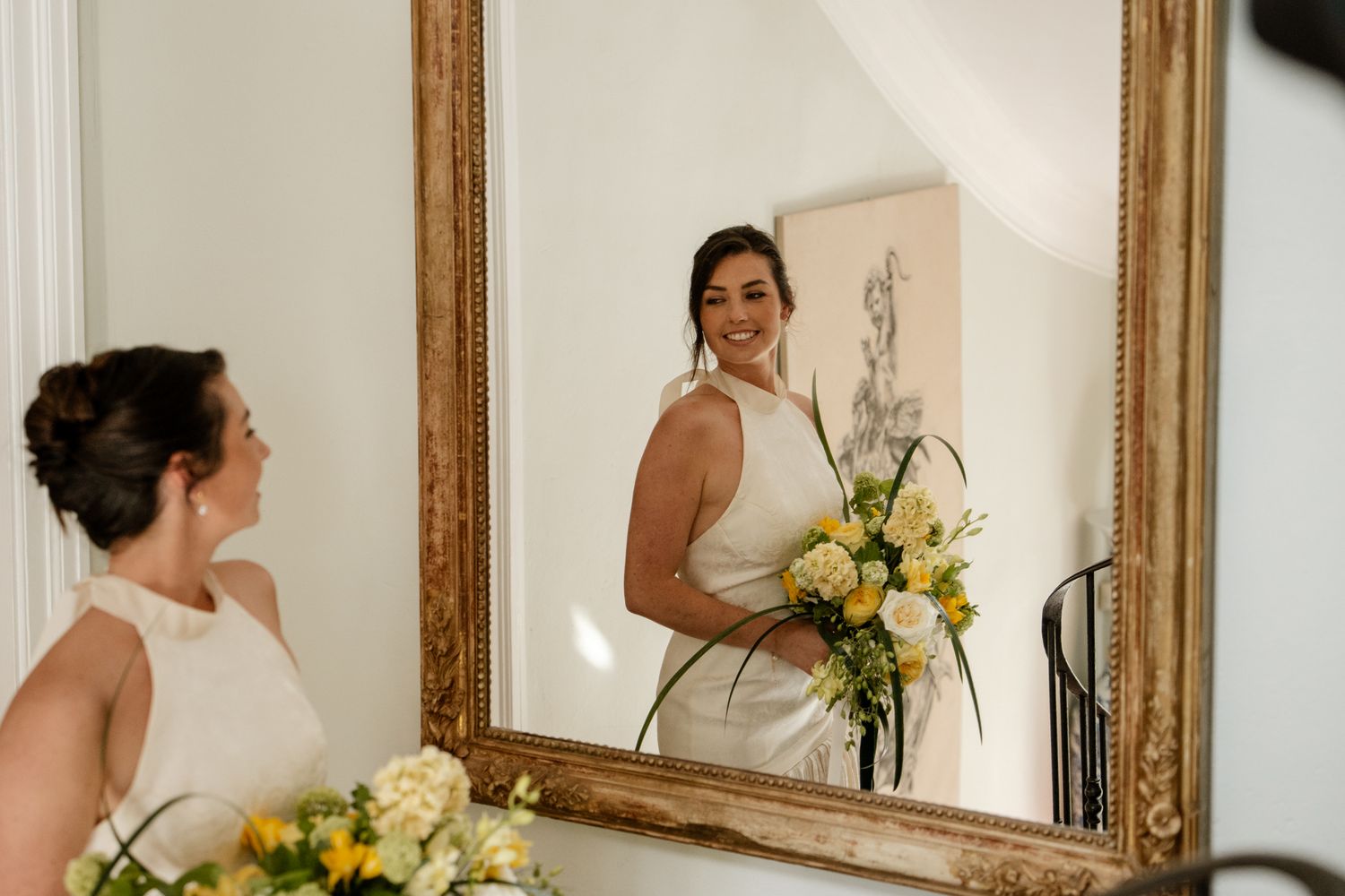 Smiling bride in white sleeveless gown holding yellow bouquet reflected in an ornate gold-framed mirror.
