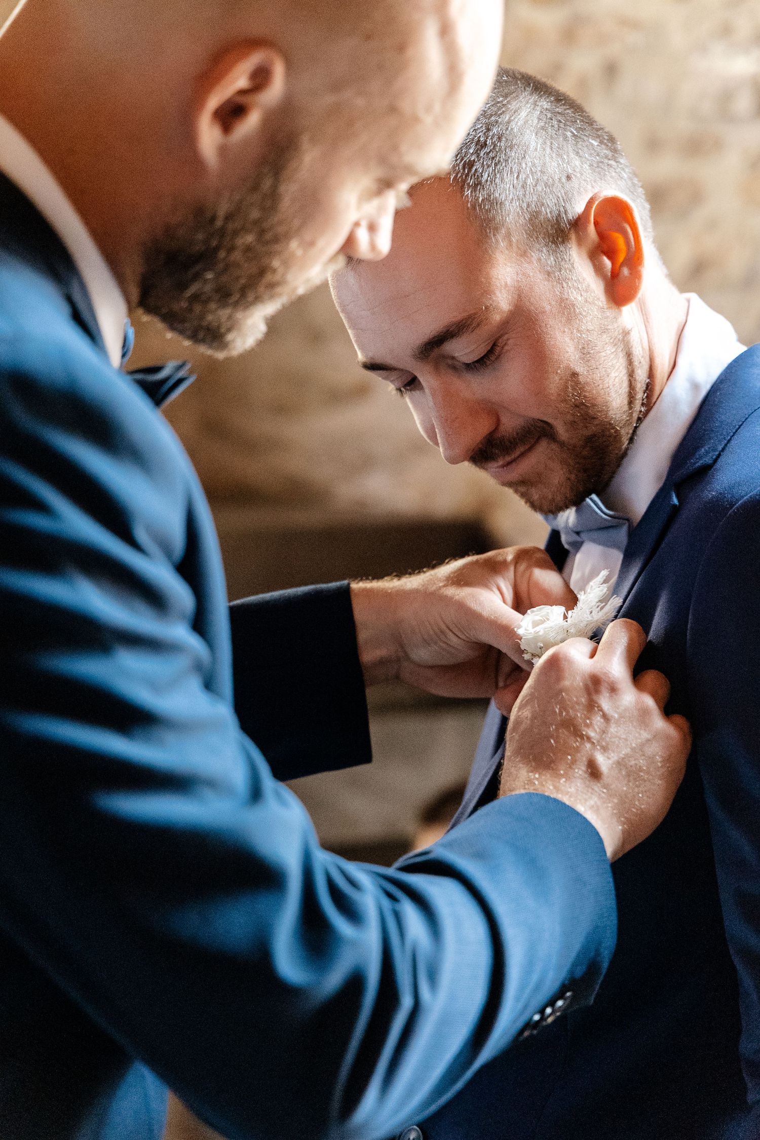 Bald groomsman carefully attaching a white floral boutonniere to a smiling man's navy suit.