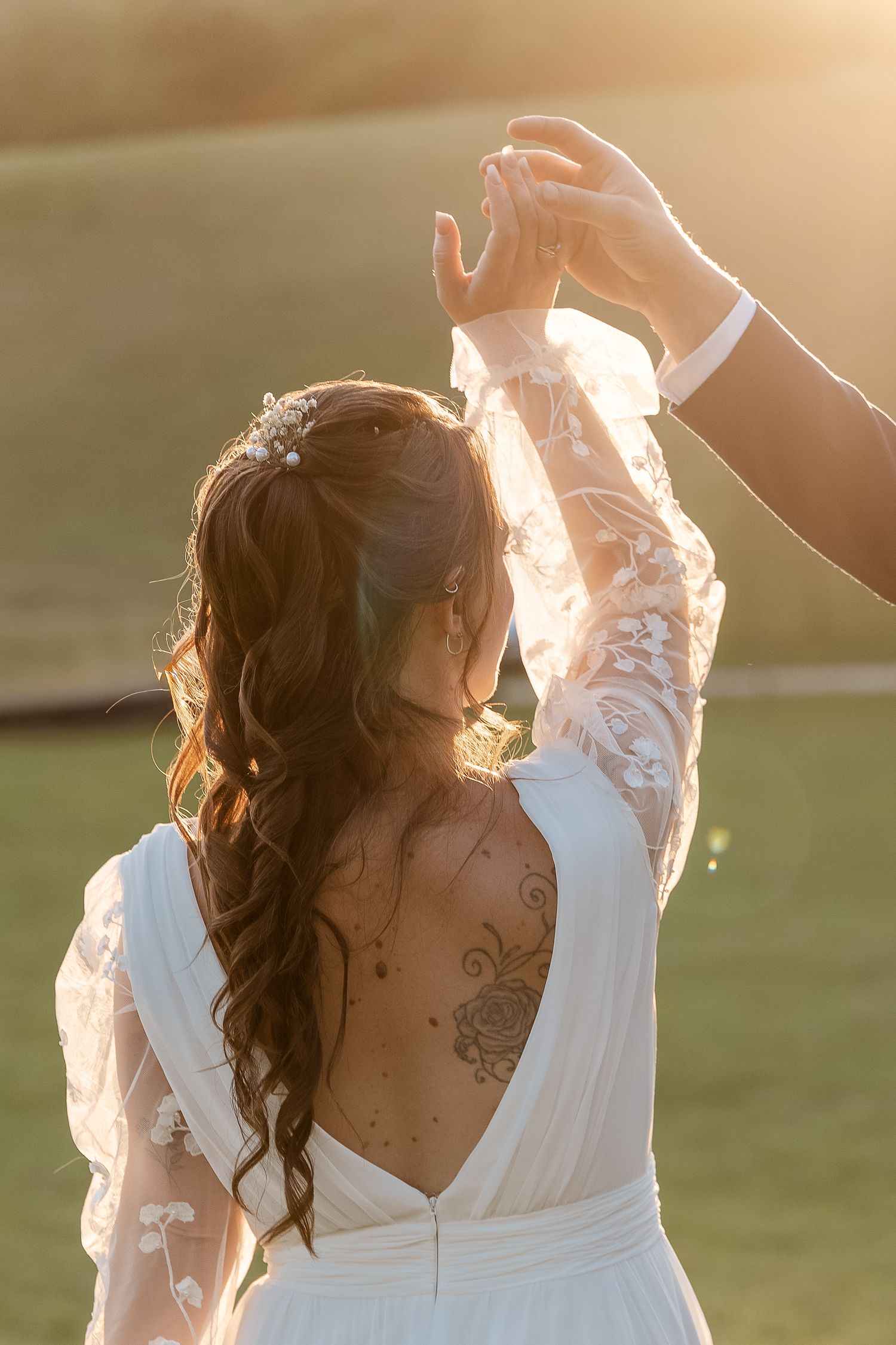 Bride with back-open lace wedding dress and tattoo raises hands with groom in warm golden sunset light.