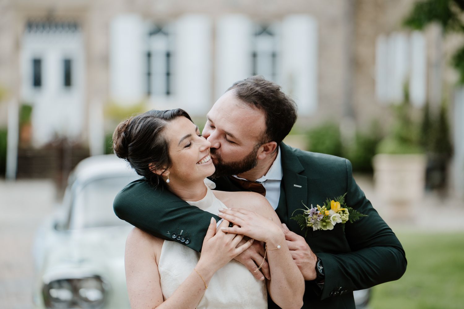 Groom in green suit kissing bride on cheek outside chateau with vintage car.