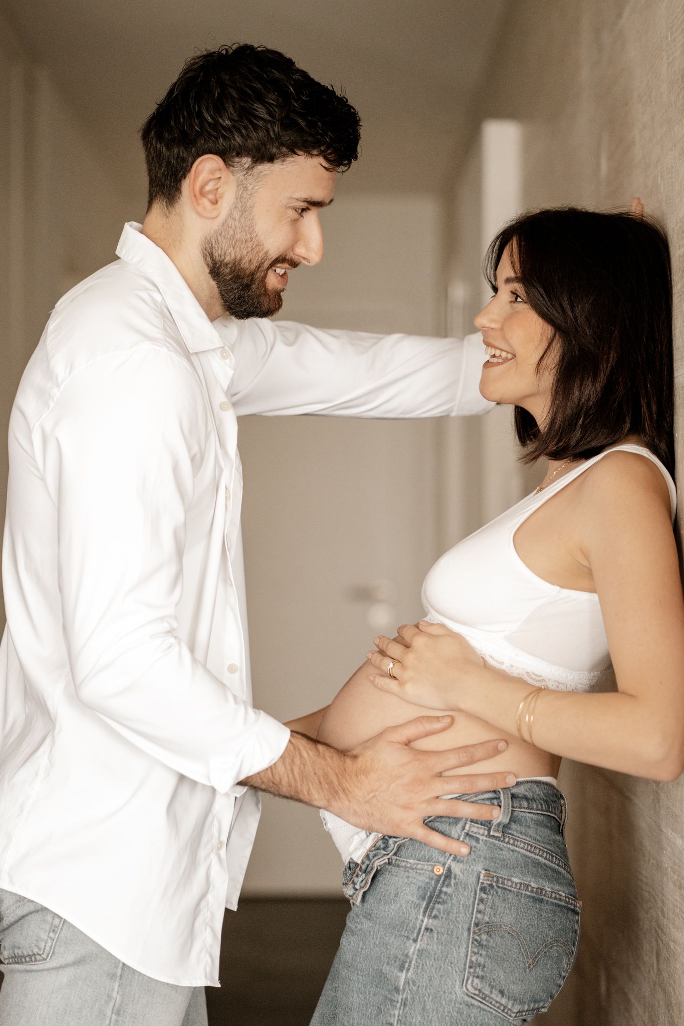 Bearded man in white shirt tenderly holding pregnant womans belly against a light wall.