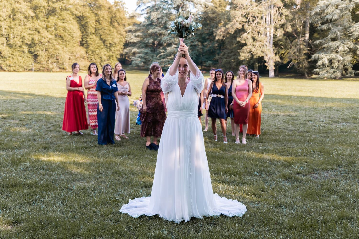 Bride in white gown tosses bouquet to group of women in colorful dresses on green lawn at outdoor wedding.