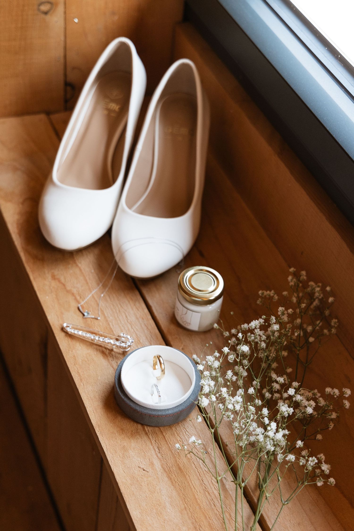 White bridal heels, wedding rings, a candle, and baby's breath flowers on a wooden shelf.
