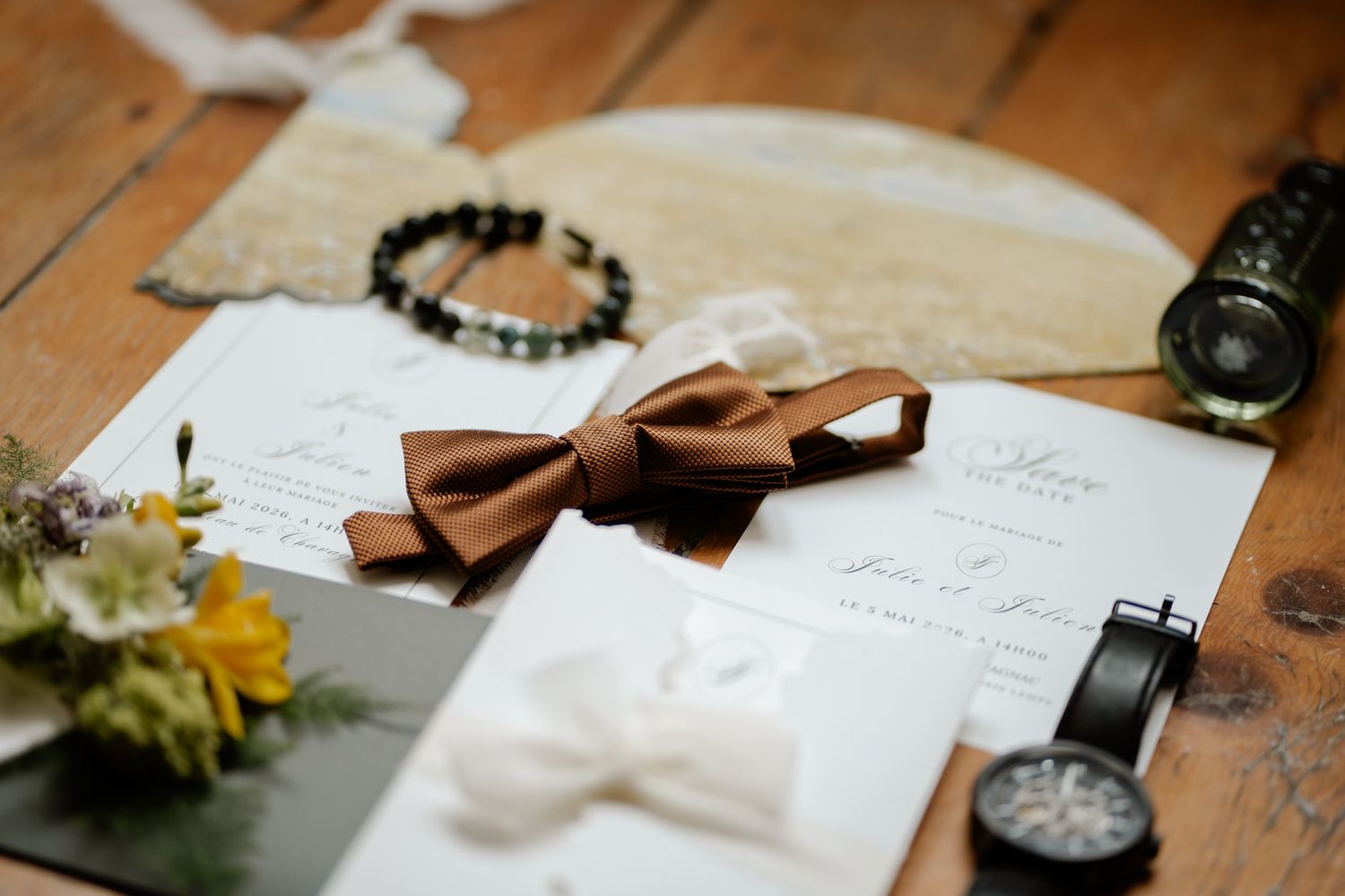 Wedding flat lay featuring brown bow tie, white invitations, watches, and bracelet on wood surface.