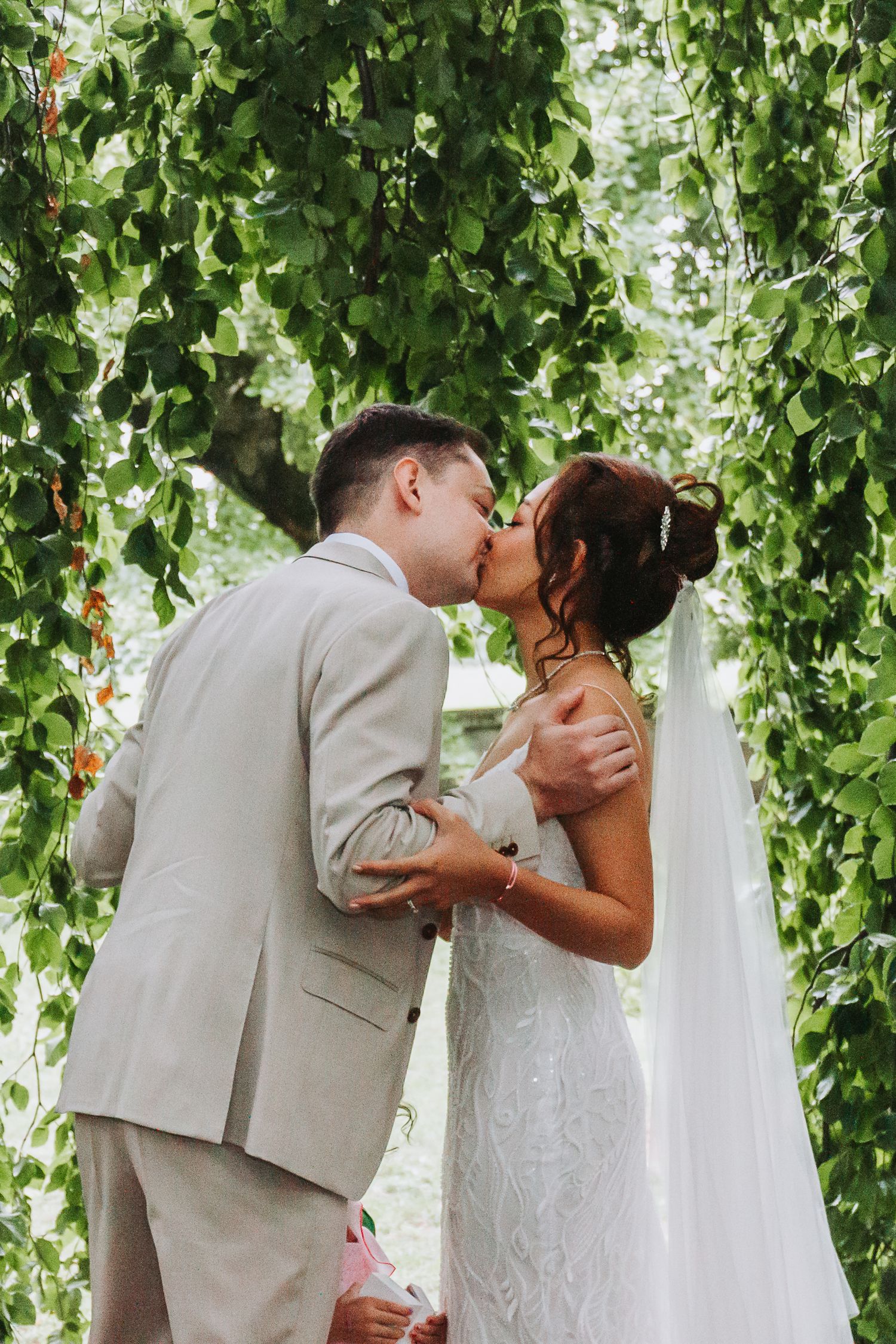 Bride and groom share their first kiss under lush cascading green branches during outdoor wedding ceremony.