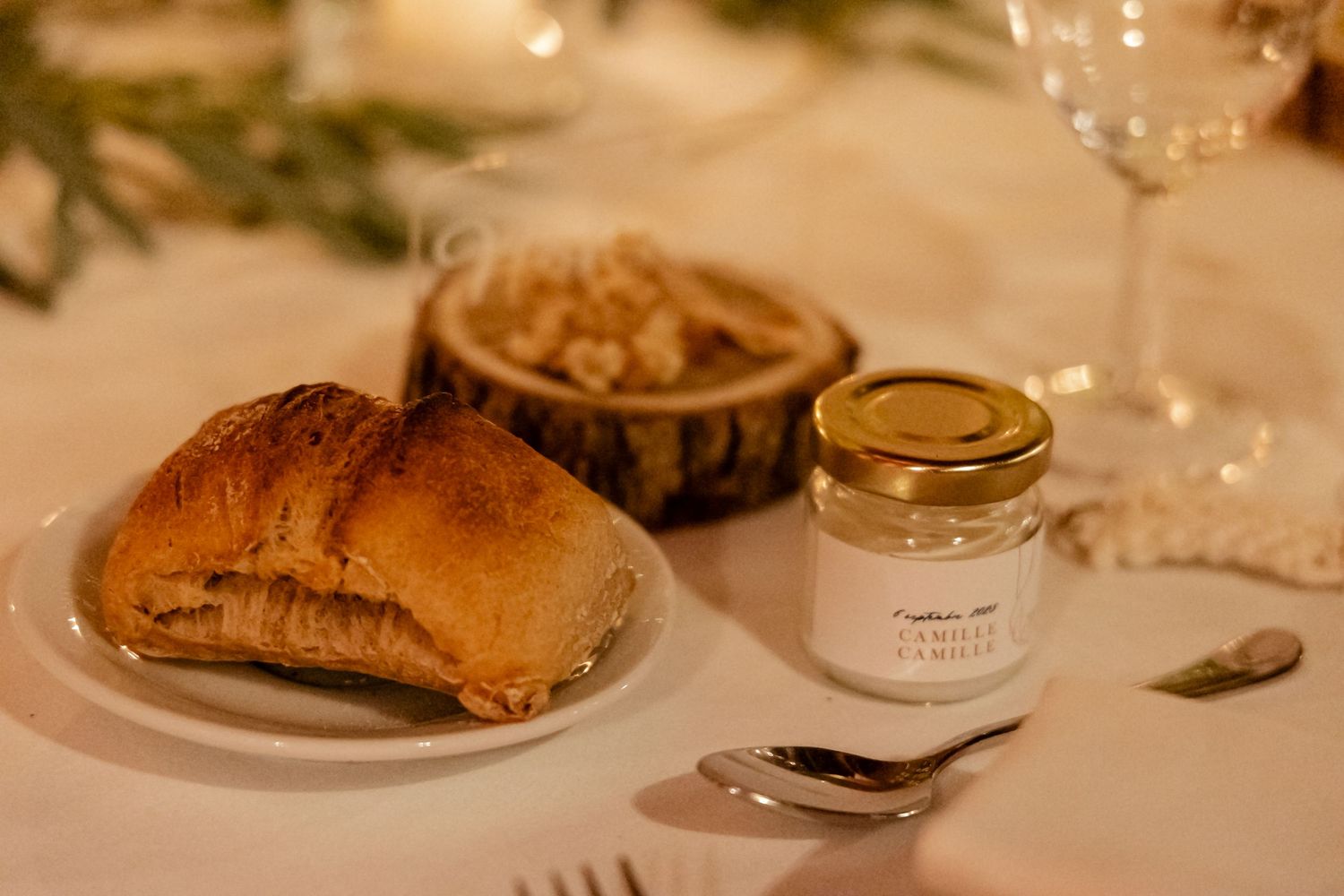 Wedding place setting with croissant, honey jar, and wine glass on white tablecloth with wood accent.
