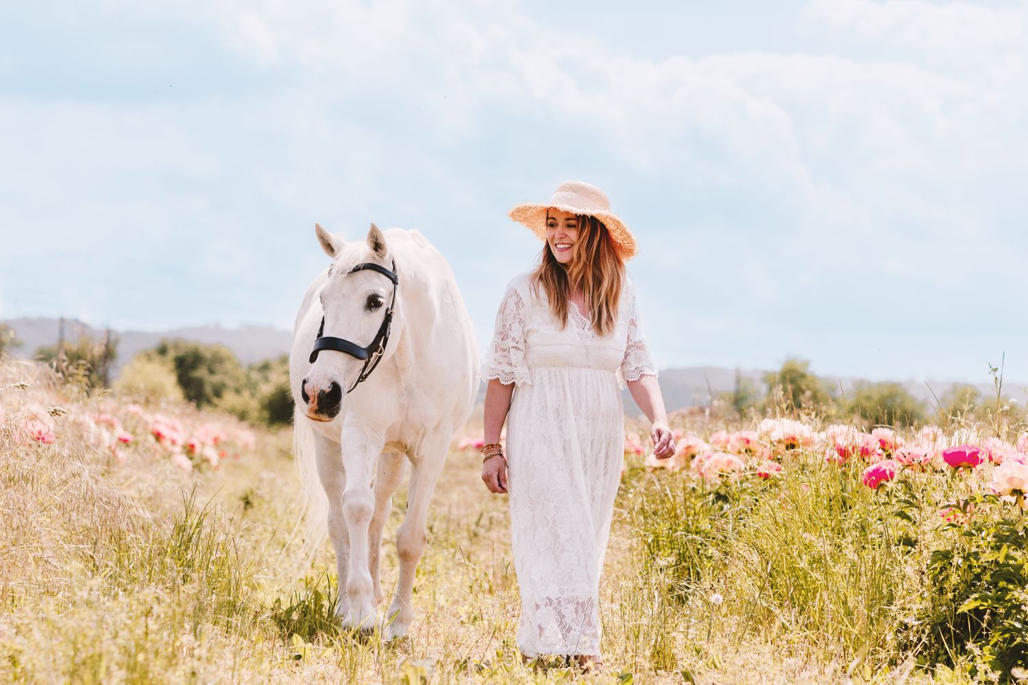 A smiling woman in a straw hat walks with a white horse through pink flowers.