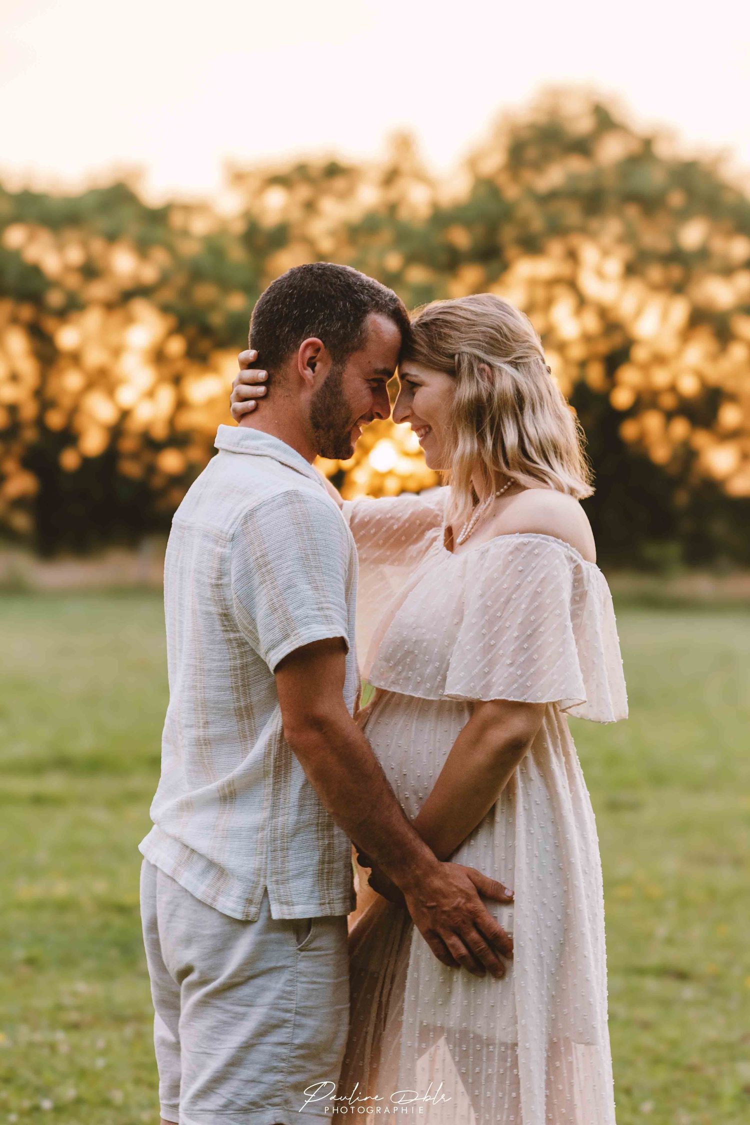 A couple shares a tender forehead-to-forehead moment in a sunlit green field at golden hour.