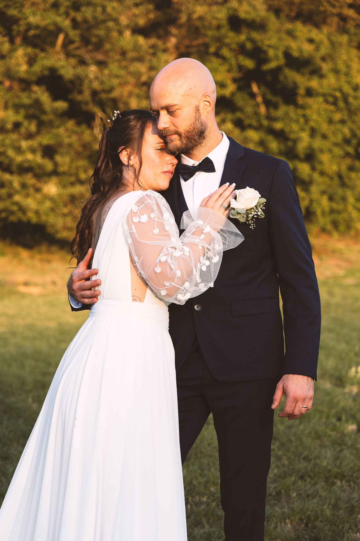 Groom in navy suit tenderly embraces bride in lace wedding dress during golden hour outdoor portrait session.