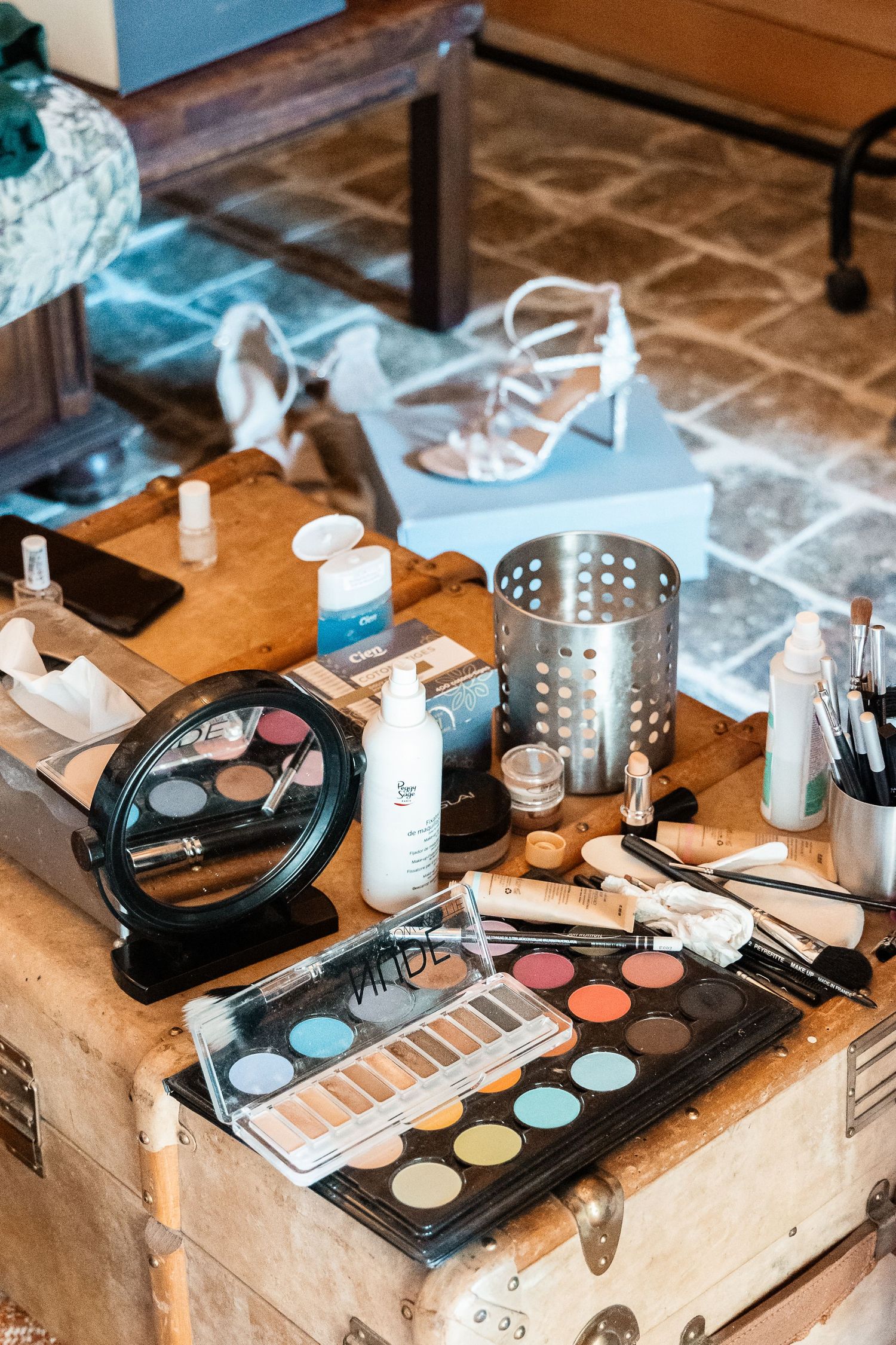 Professional makeup artist tools including eyeshadow palettes, brushes, and skincare products on a vintage wooden trunk.