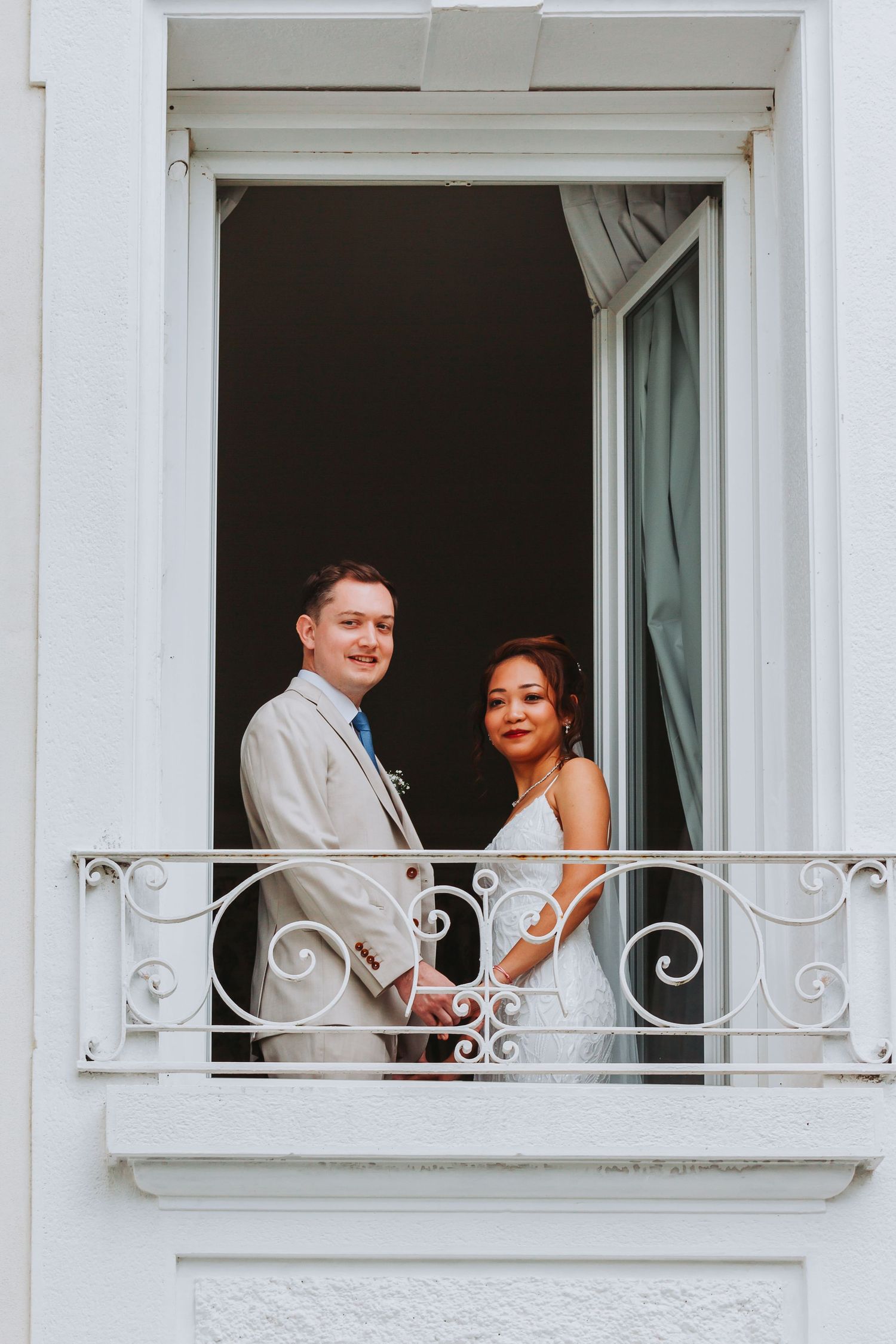 Smiling couple holding hands on a white ornate balcony, groom in beige suit and bride in white dress.