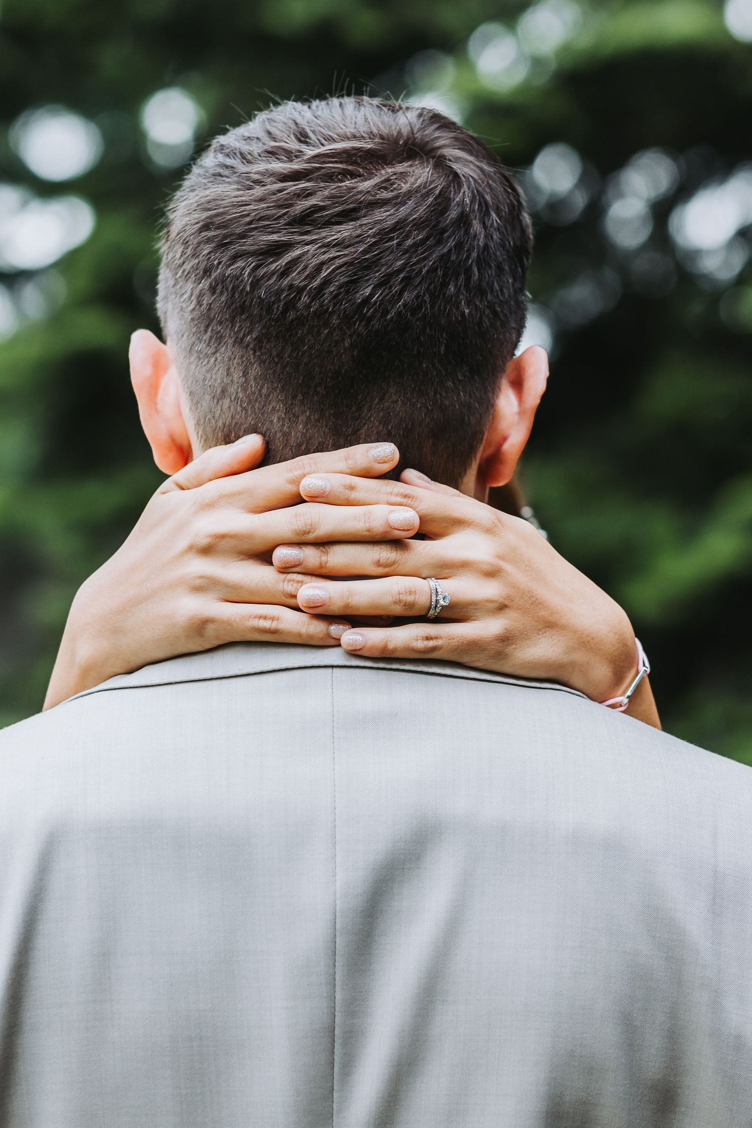 Back view of bride's hands clasped around groom's neck, wedding ring visible, groom in light gray suit.