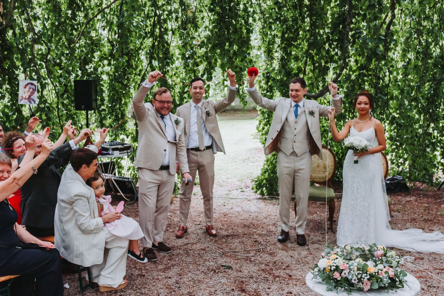 Joyful wedding ceremony outdoors as bride, groom and groomsmen raise glasses in celebration under lush green hanging foliage.