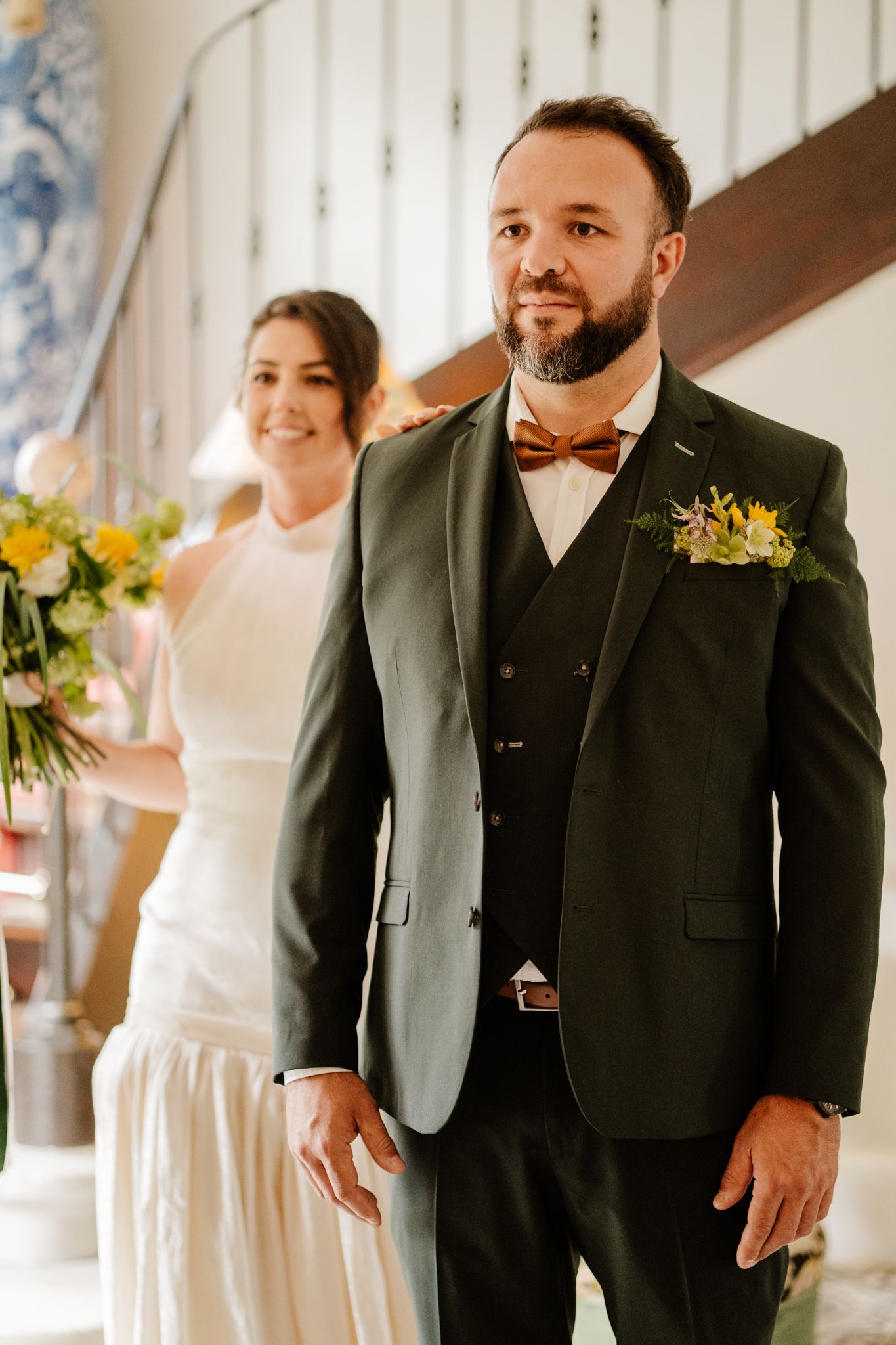 Groom in green three-piece suit with wooden bow tie and floral boutonniere standing with bride.