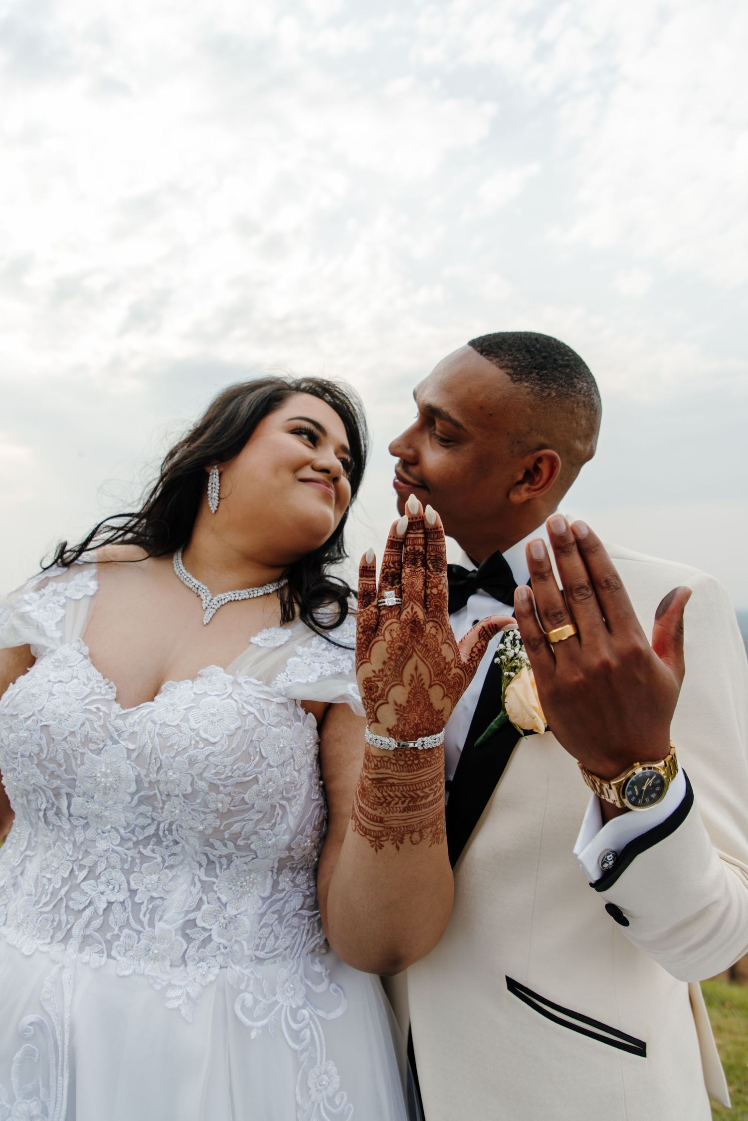 Newlywed couple showing off their wedding rings and henna decorated hands against cloudy sky.