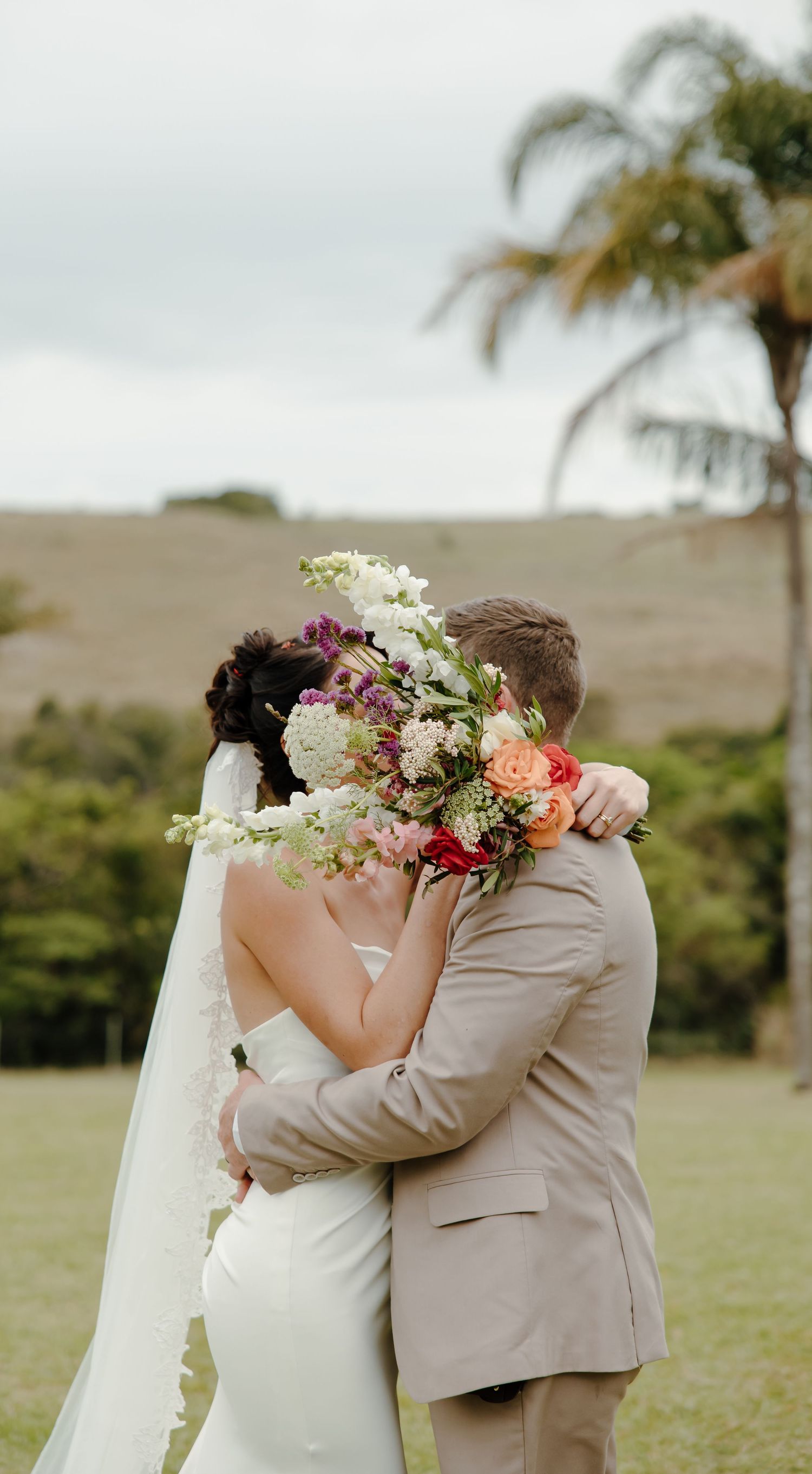 A wedding couple embraces behind a colorful bouquet with a palm tree and rolling hills in the background.