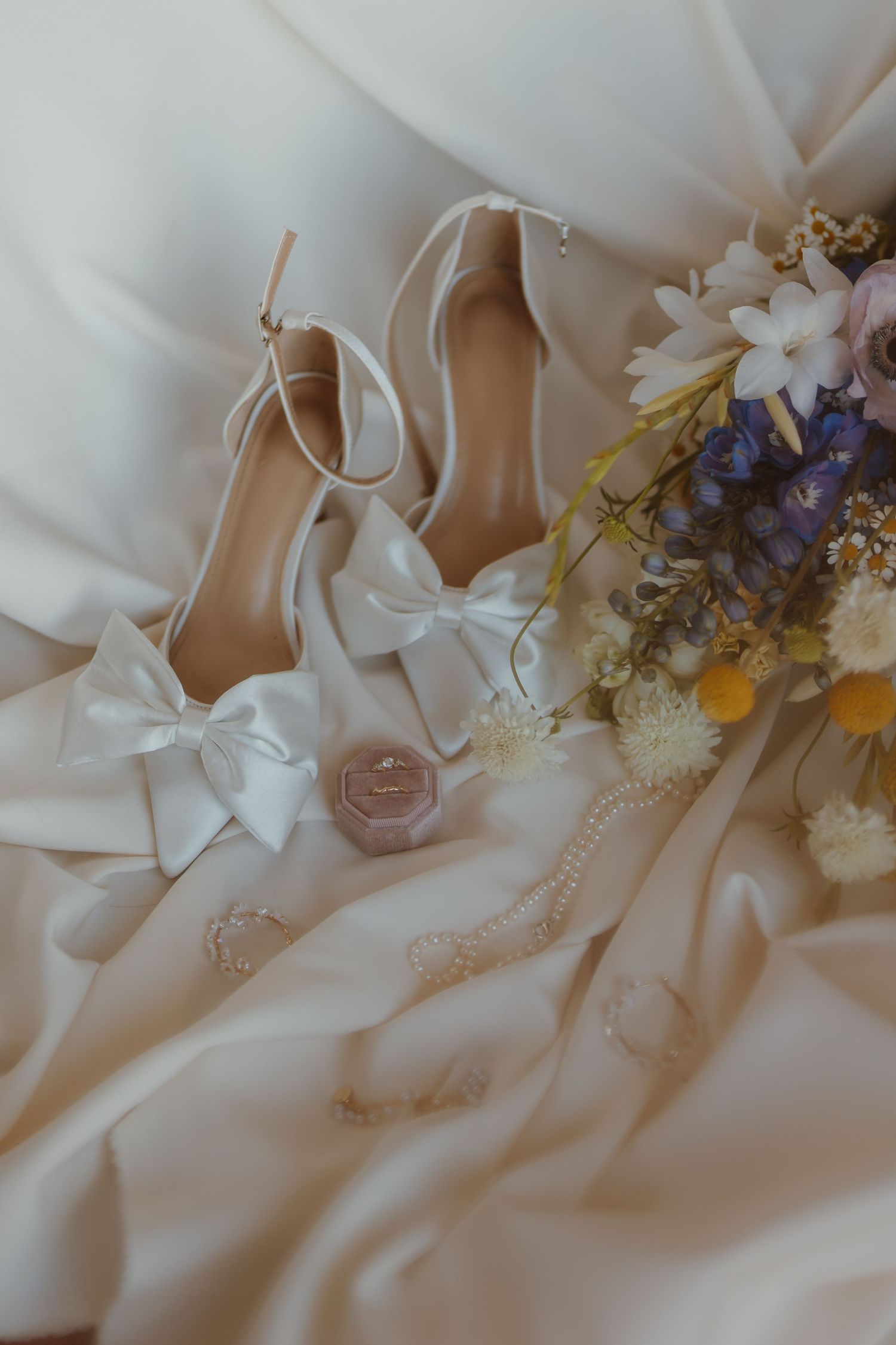 White wedding shoes with delicate bows arranged with flowers and pearls on flowing ivory fabric.