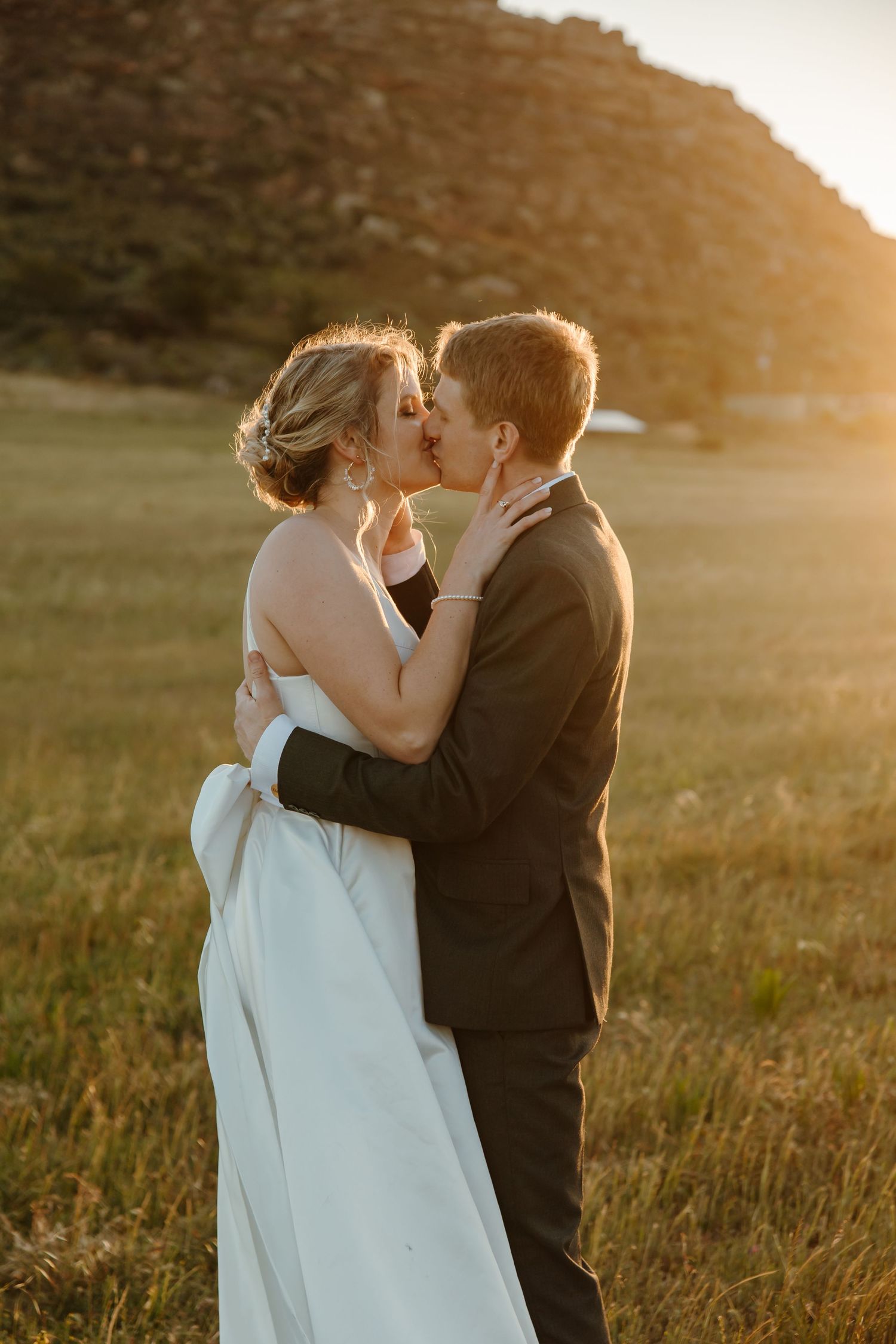 A couple shares a romantic sunset kiss in a golden field wearing wedding attire.
