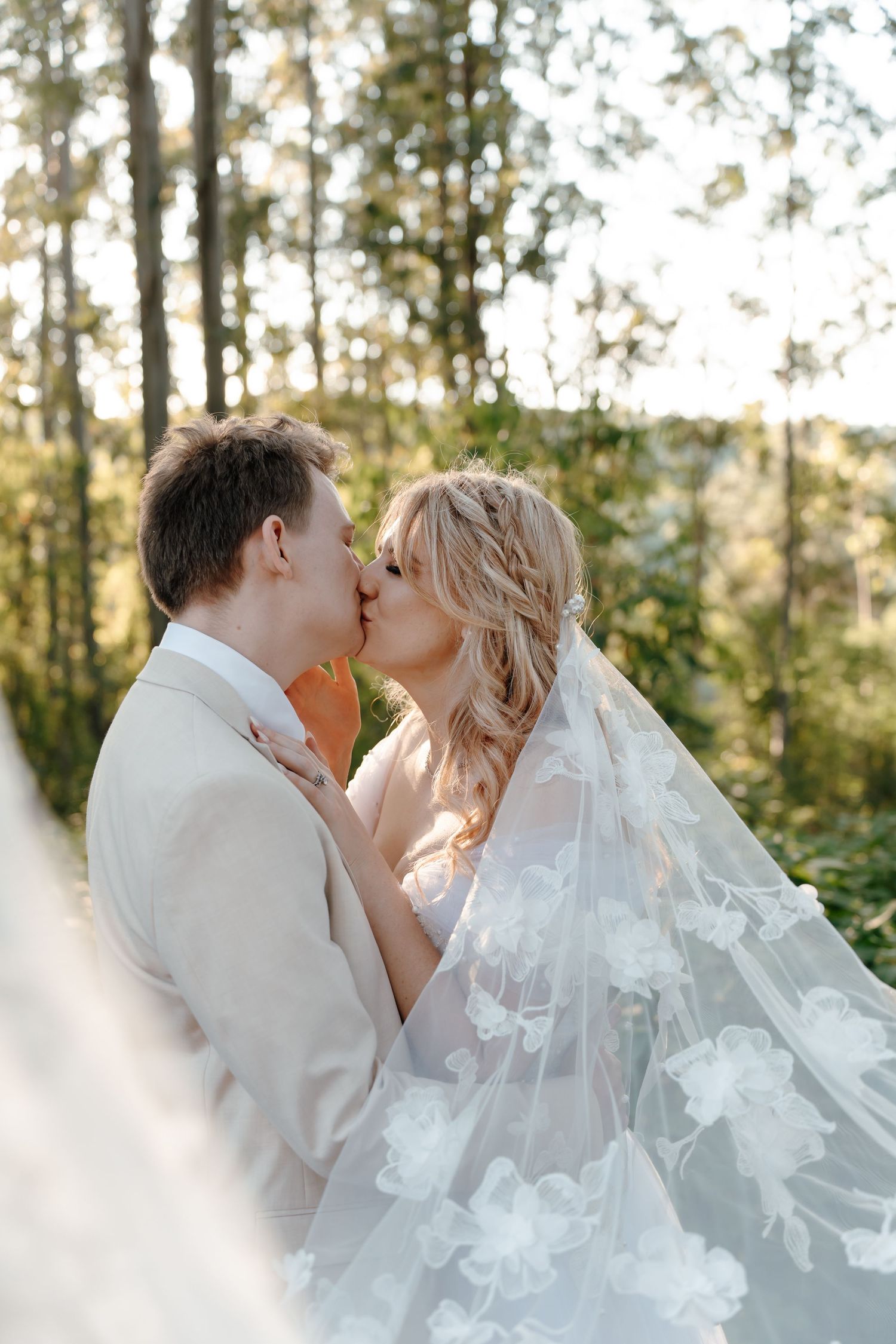 A romantic wedding couple shares a tender kiss outdoors with sunlit trees and a flowing white veil in the background.