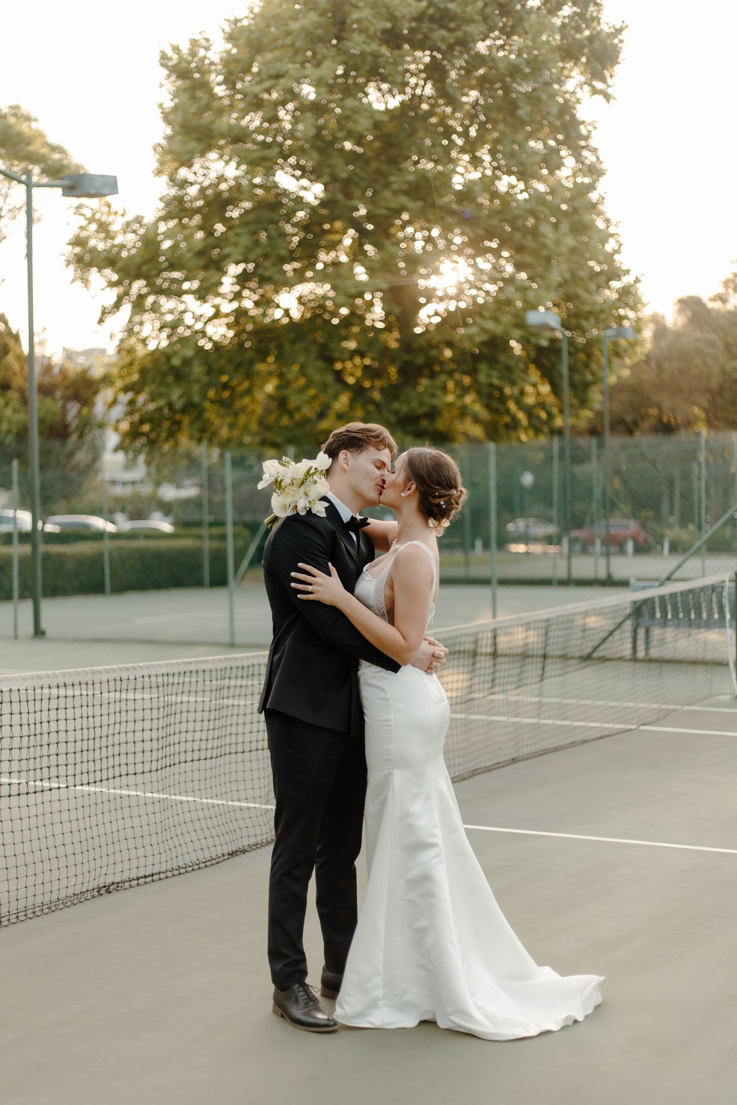 Romantic couple in wedding attire embrace on outdoor tennis court at sunset.