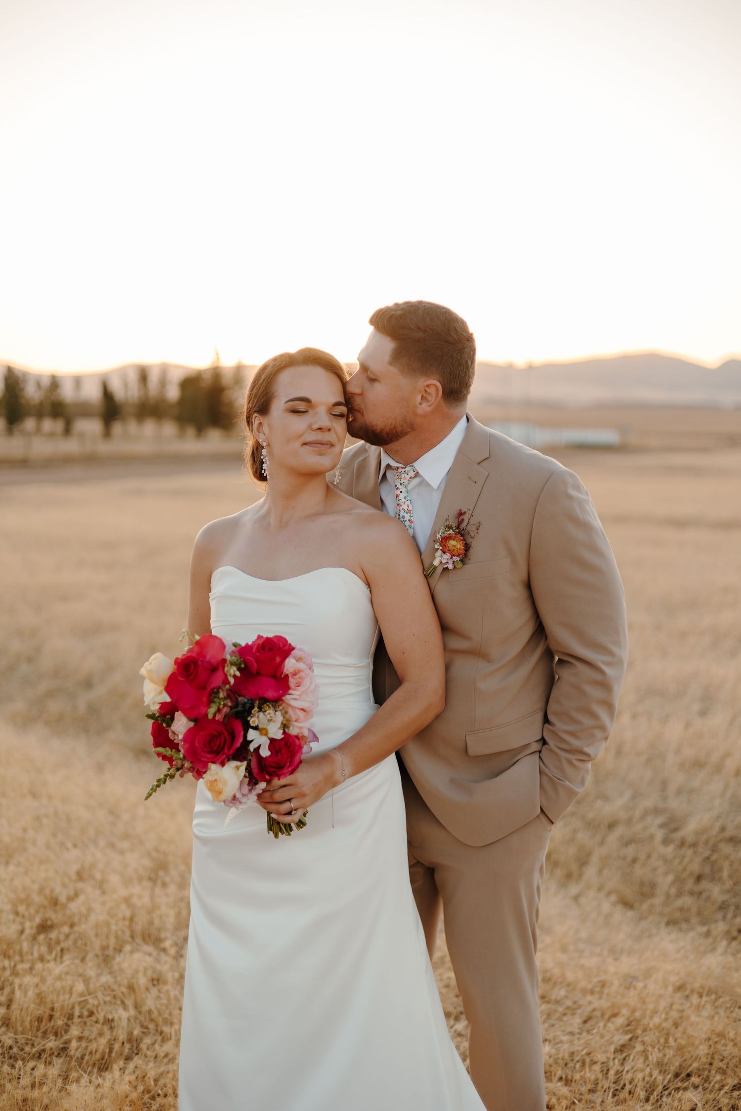 A sunset wedding portrait in a golden field with vibrant red bouquet.