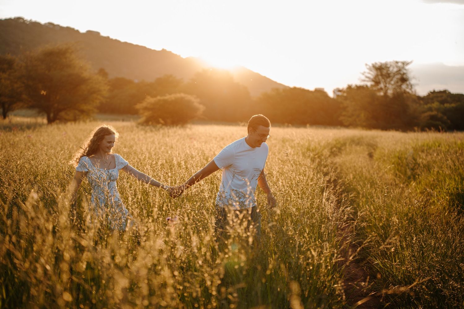 Couple walking through golden wheat field at sunset holding hands in romantic summer countryside setting.