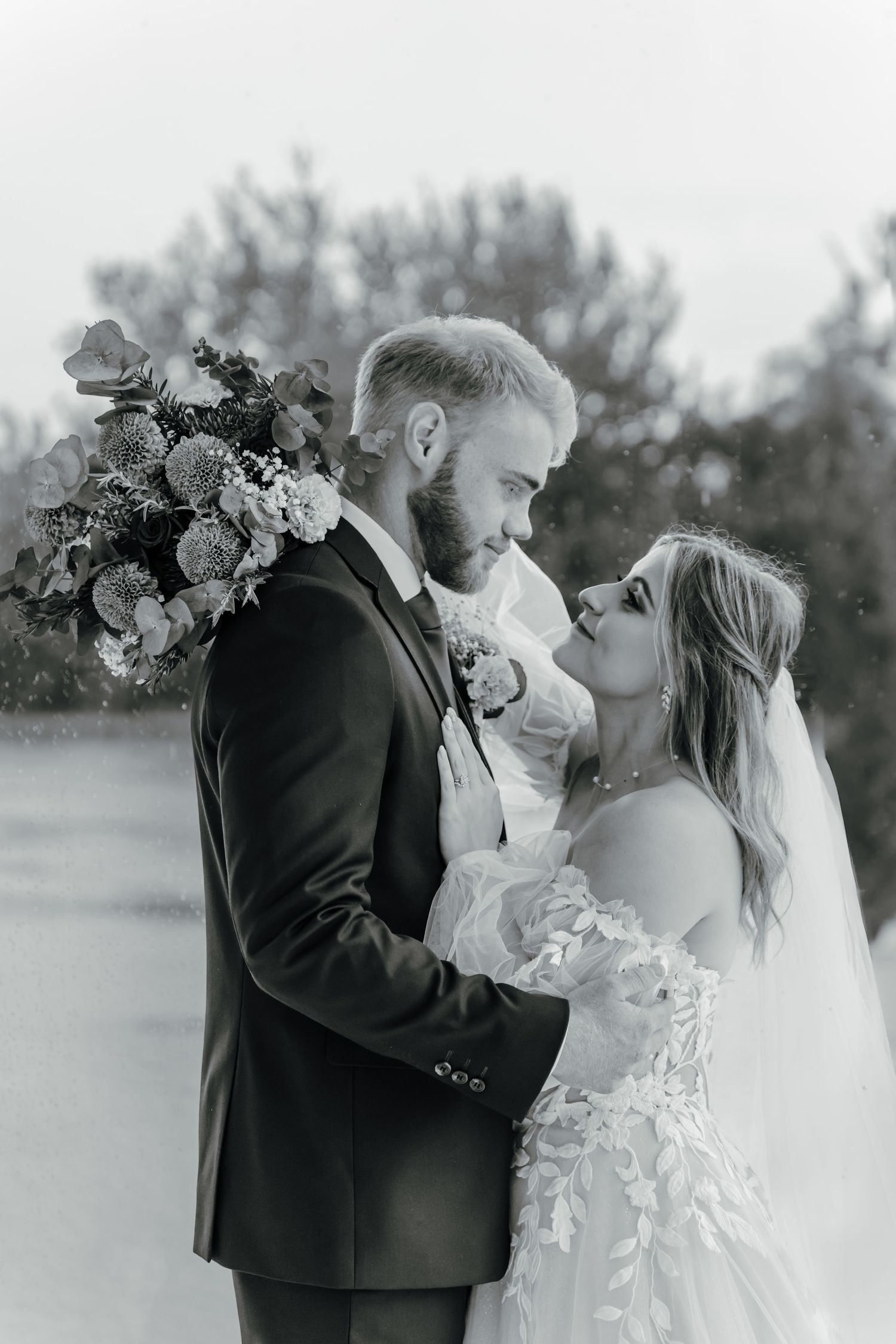 A romantic black and white wedding photo of a couple embracing outdoors with a beautiful floral bouquet.