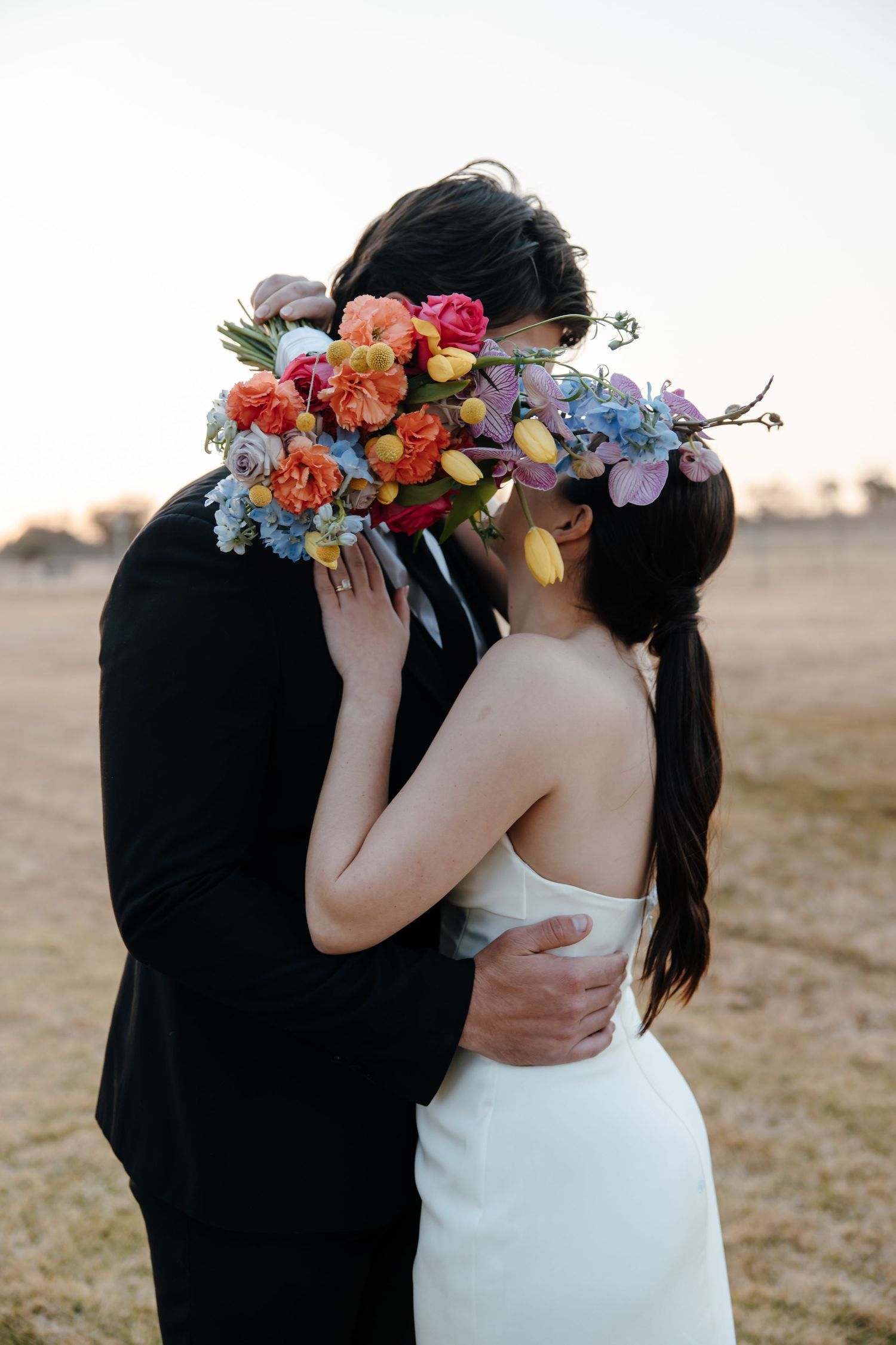 Romantic wedding couple embraces in a field at sunset with the bride wearing a colorful floral crown.