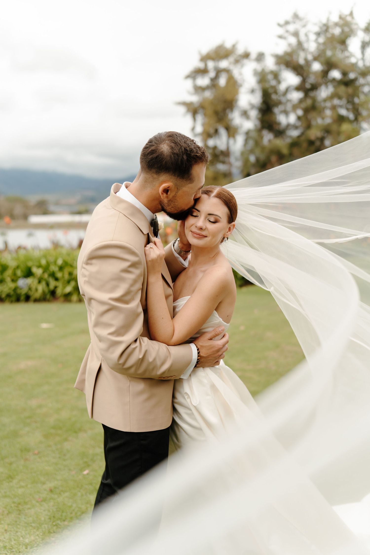 A romantic wedding couple embraces on a grassy field while a flowing white veil creates an ethereal effect in the breeze.