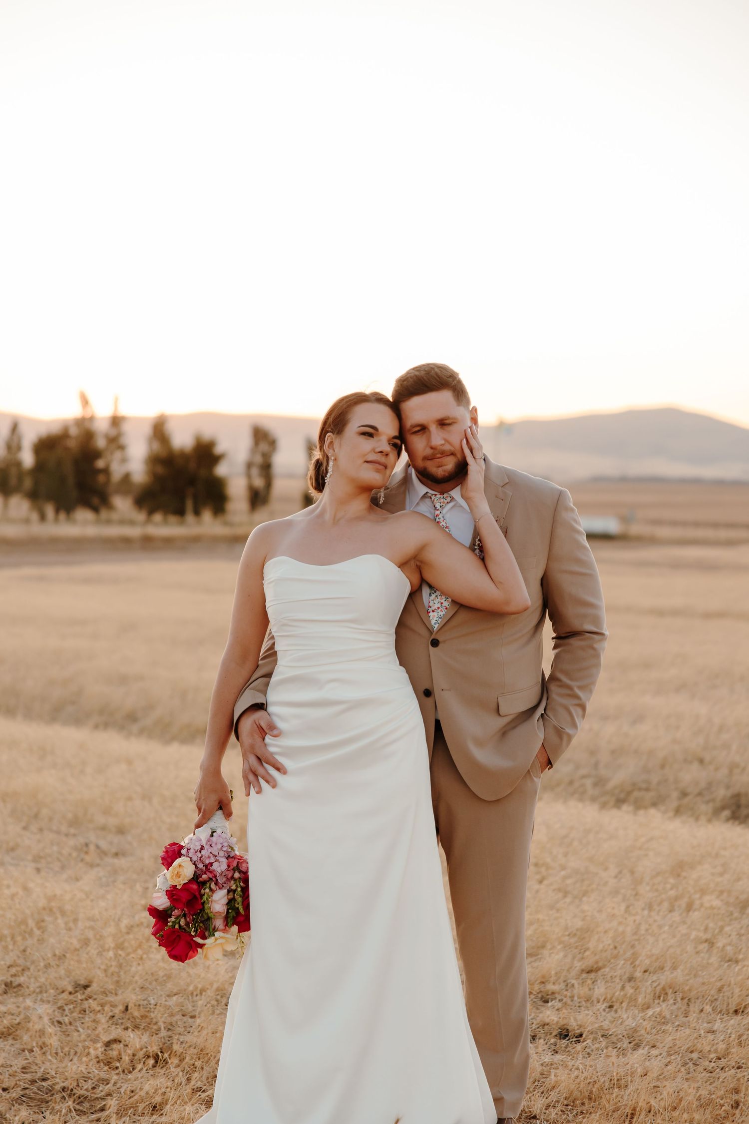 A couple embraces in a golden wheat field during sunset while wearing wedding attire.