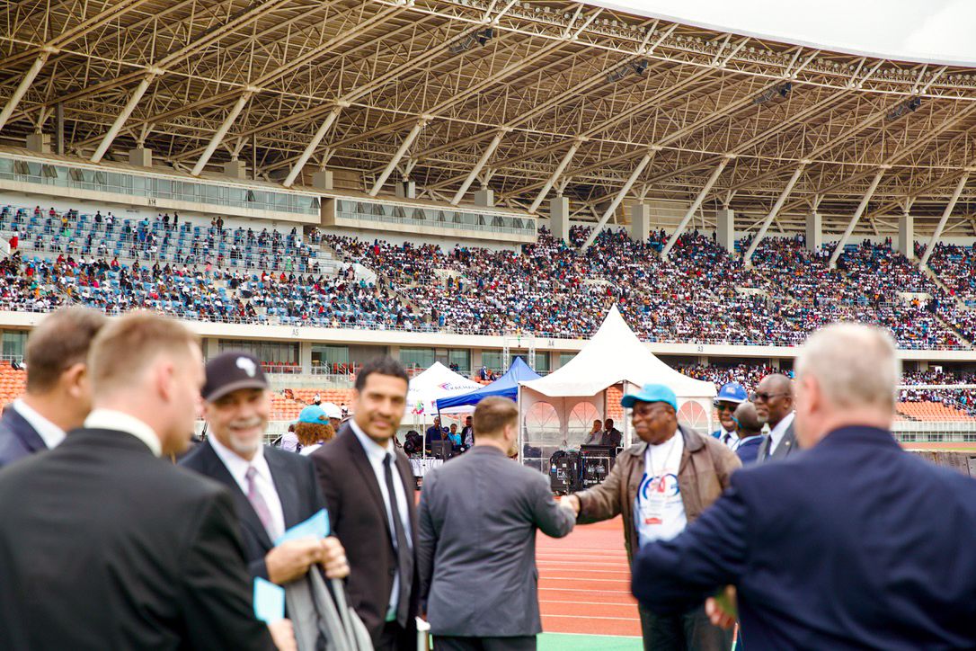 Dignitaries in suits greeting each other on a stadium track at a packed sports event in Africa.