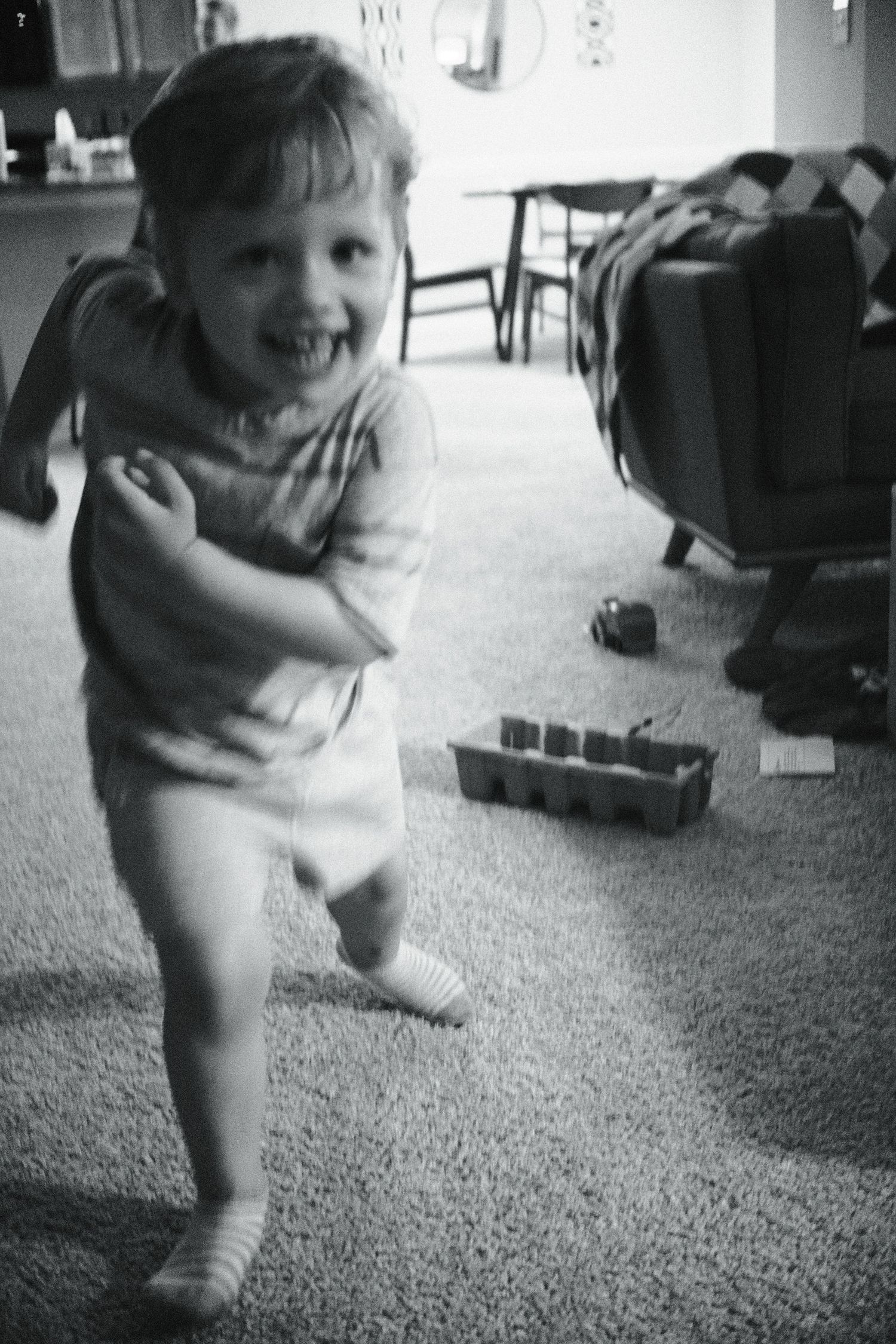 Happy toddler girl running and smiling indoors in a black and white candid photo.