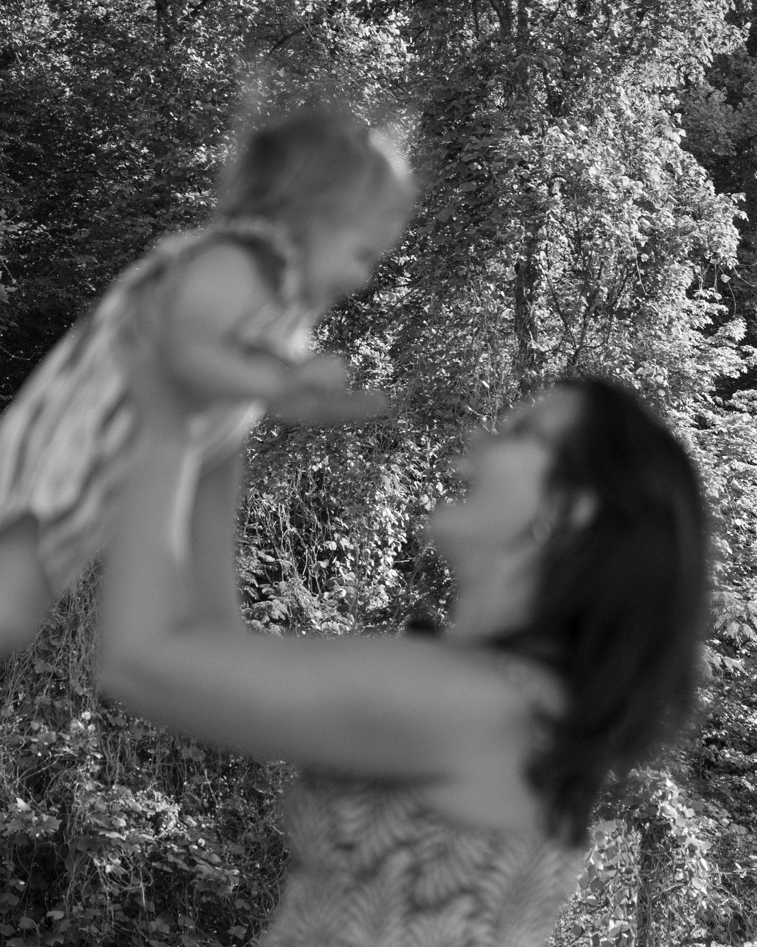 Black and white photo of mother lifting baby daughter up among leafy trees.