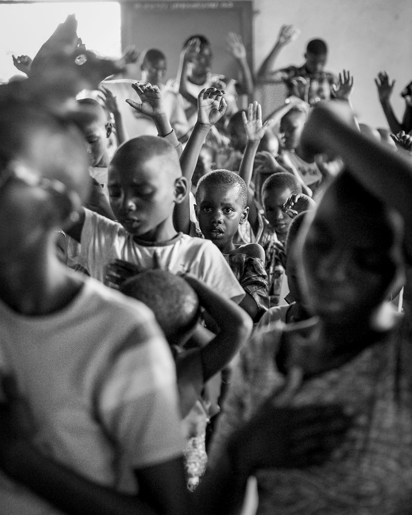 Black and white photo of African children raising hands in a crowded indoor gathering.