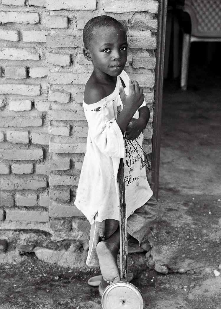 Young African child in white dress standing beside scooter toy near brick wall, black and white.