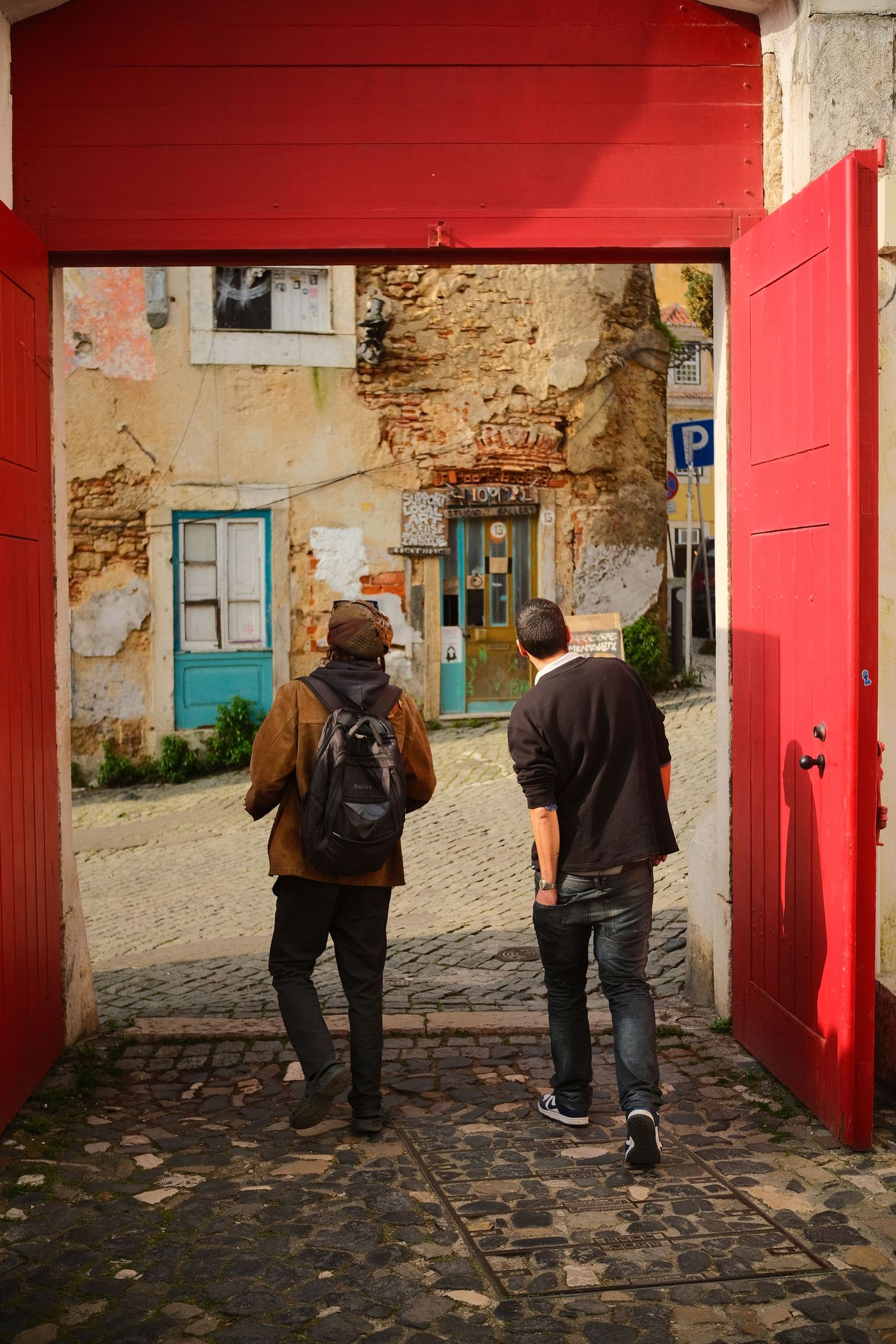 Two men walking through a red gate into a rustic cobblestone alley in Lisbon, Portugal.