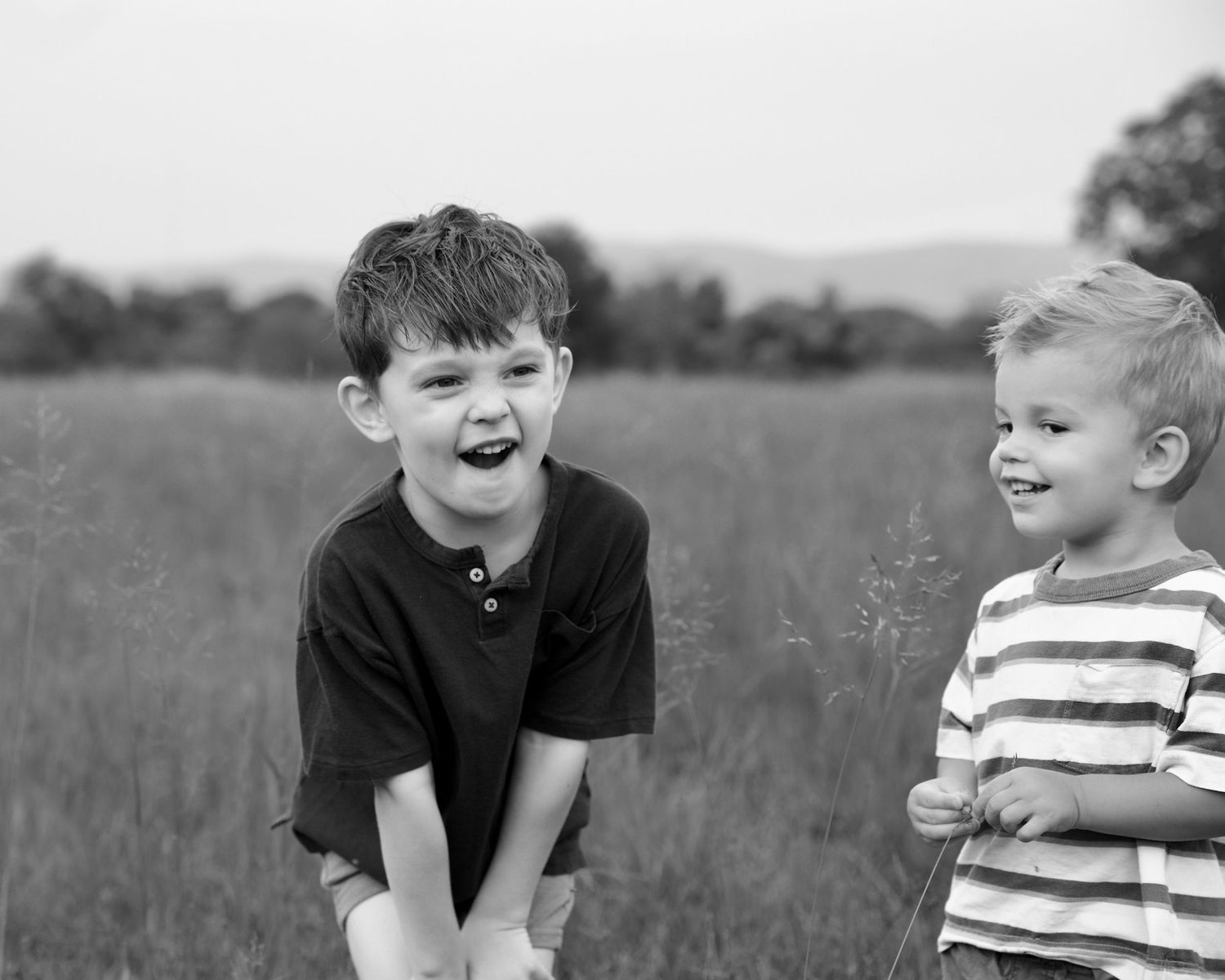 Two young boys laugh and smile together in a black and white photo taken in an open grassy field.
