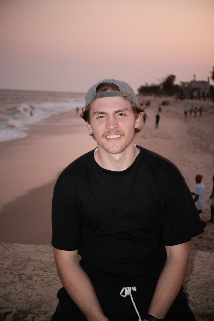Young man in a black shirt and cap smiling on a sandy beach at sunset.