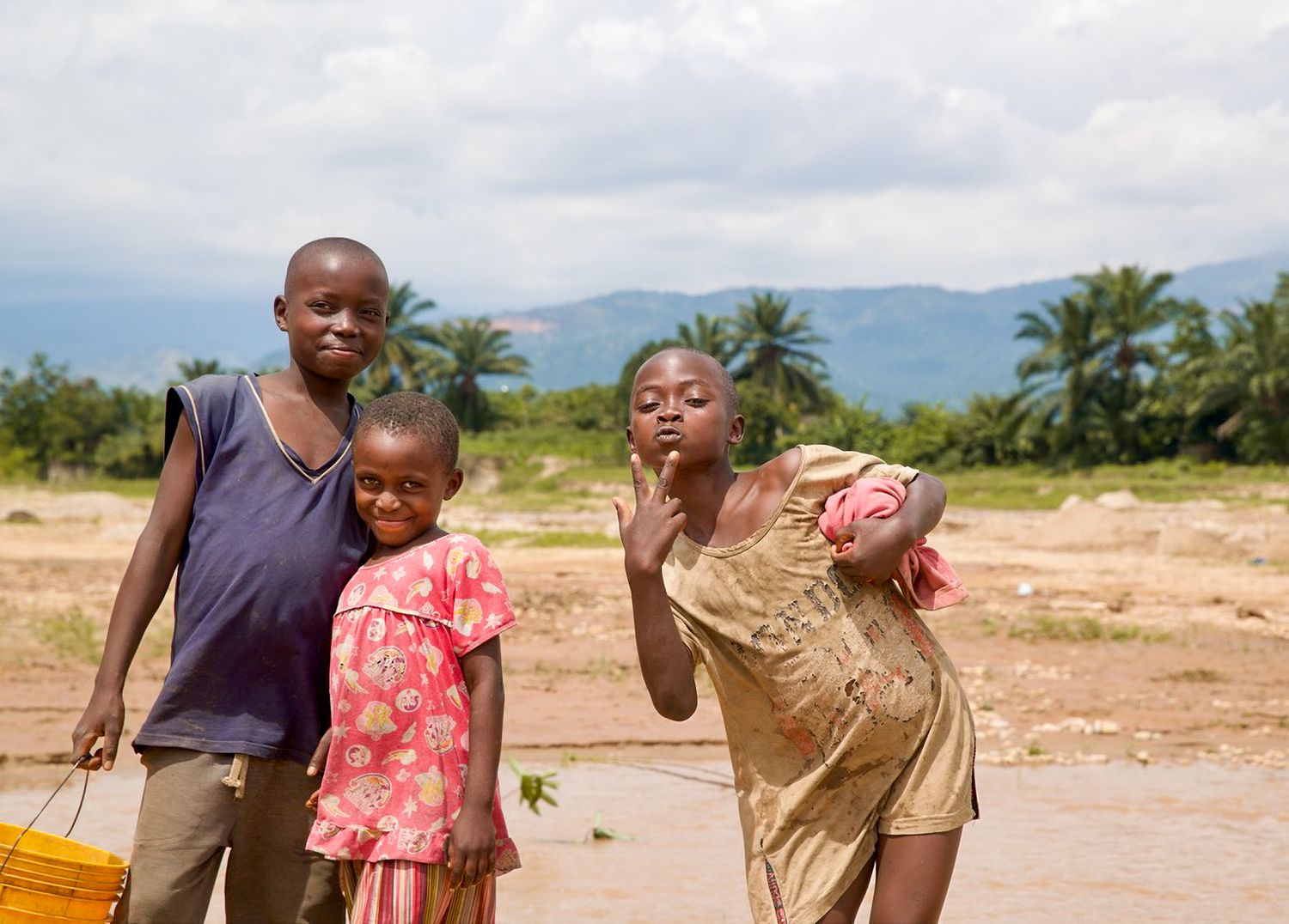 Three African children standing outdoors near a river with mountains and palm trees in background.
