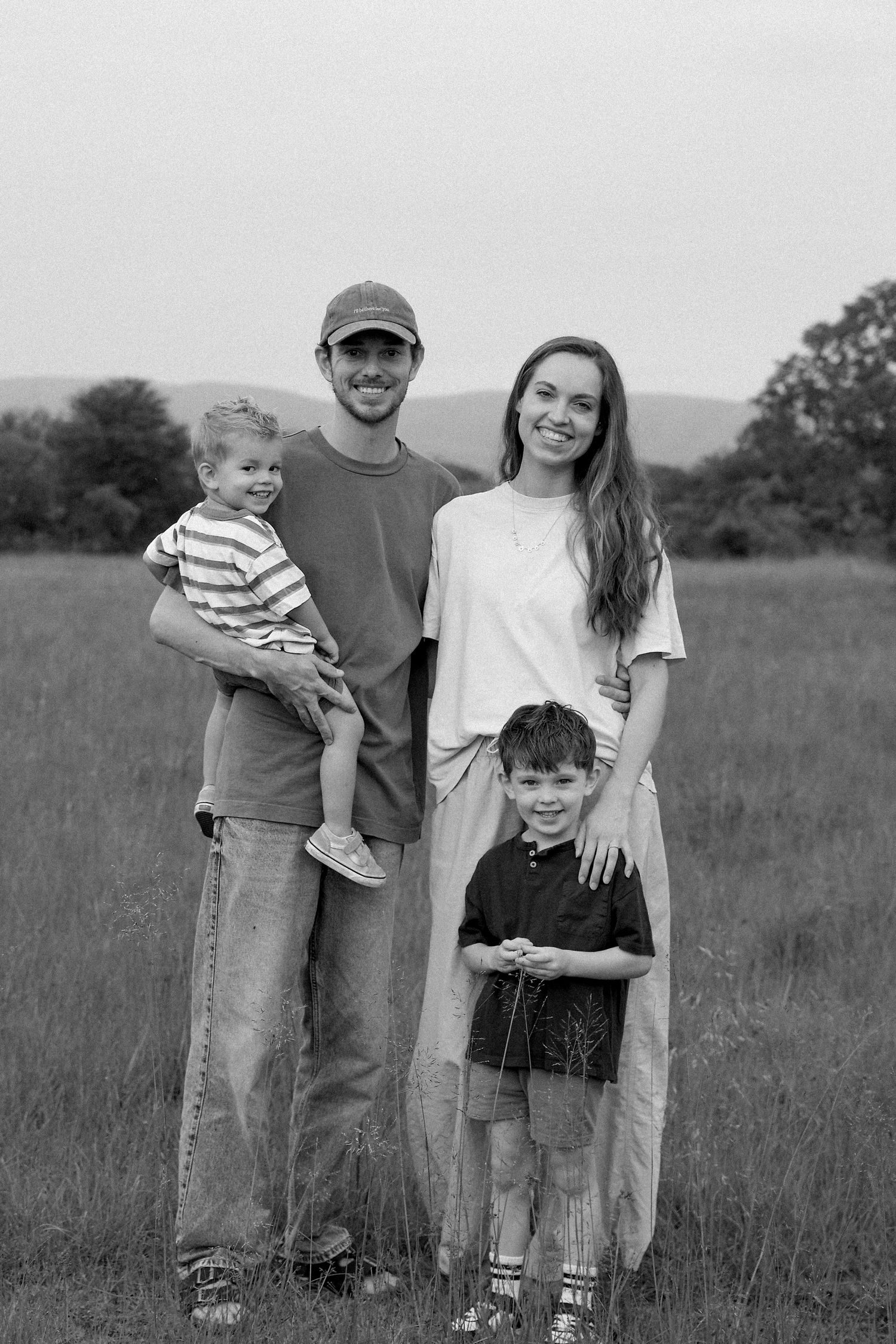 Black and white family photo of couple with two boys smiling in grassy meadow.