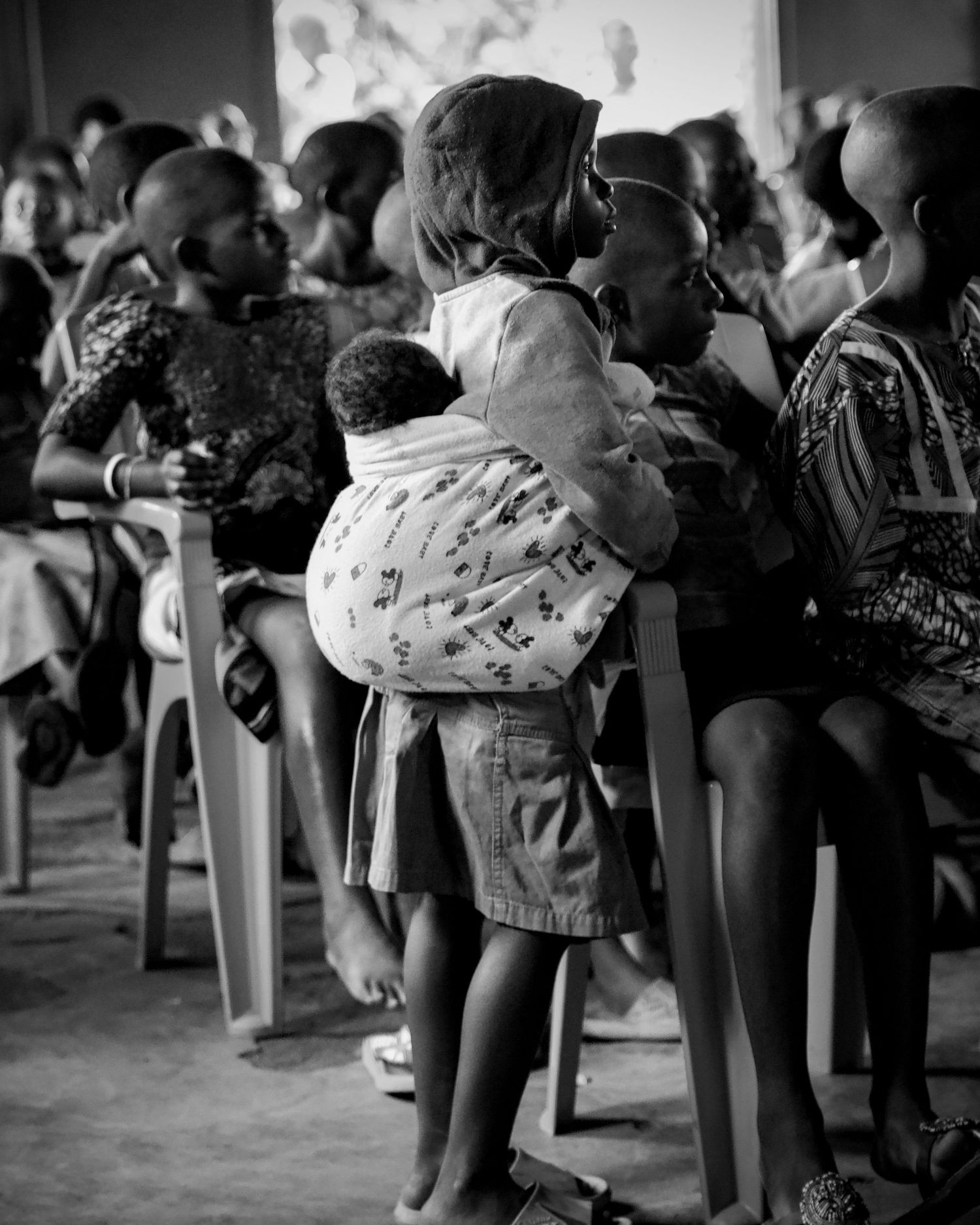 Young African girl carrying a baby on her back while seated among children in black and white.