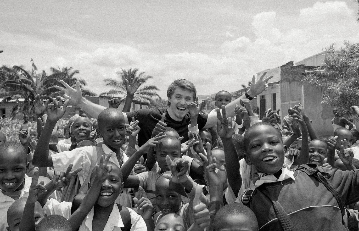 A white man surrounded by joyful African children raising their hands outdoors.