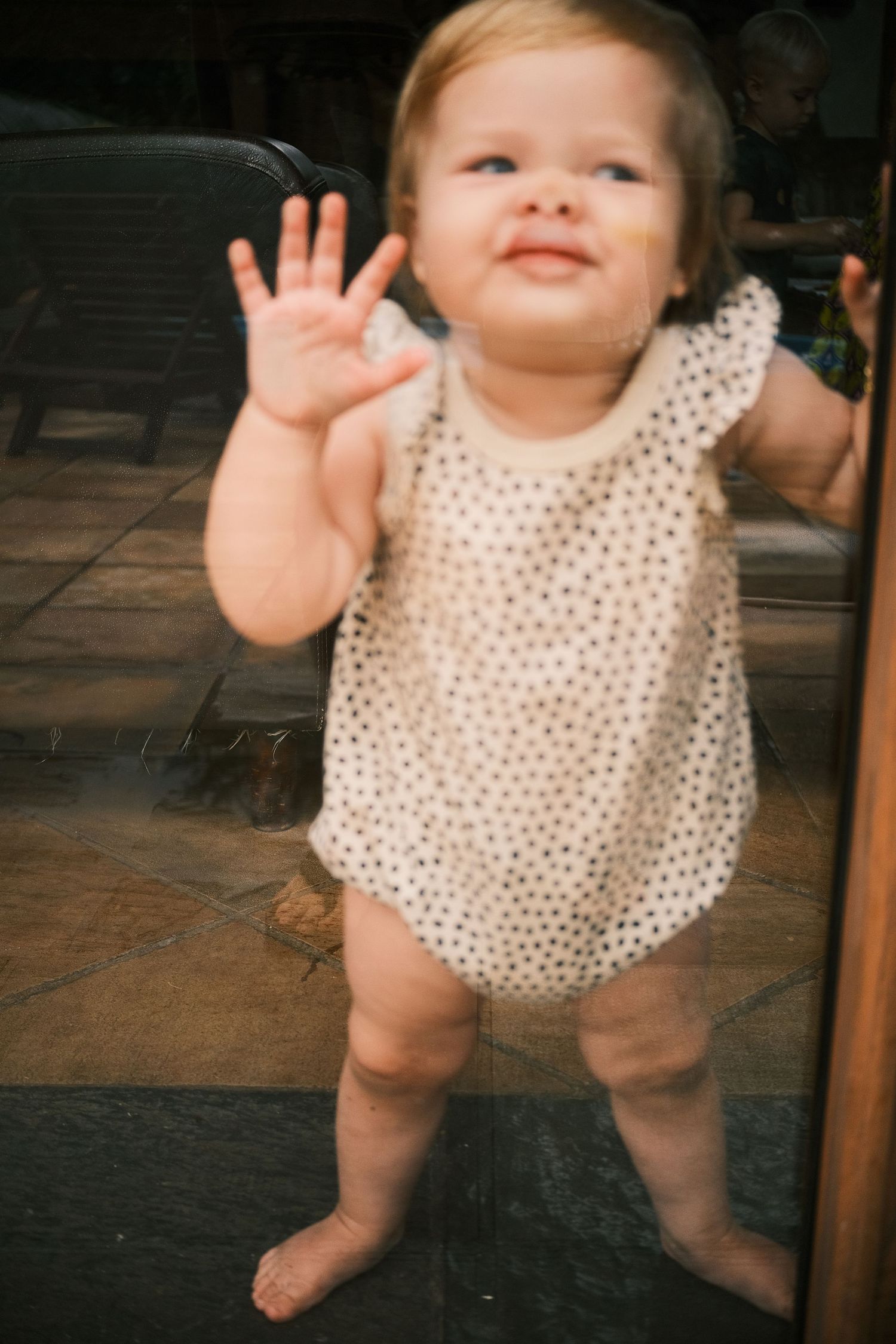 Adorable toddler in polka dot romper pressing hand against glass door while standing barefoot.